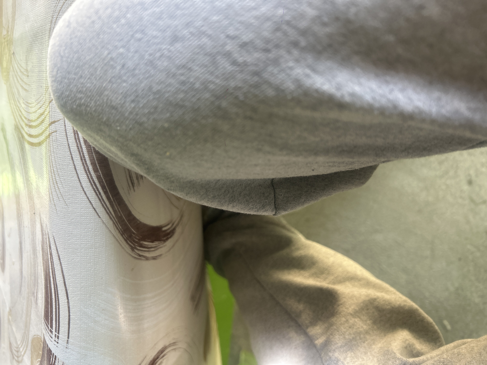 The image depicts a close-up, angled view of a person sitting at a table, likely indoors. The person is wearing light gray sweatpants. The fabric of the pants is visible in detail. A table covered with a patterned, light-colored material is to the left of the pants. The pattern on the table appears to be dark brushstrokes on a lighter background. Behind the pants and table, a section of green and light gray/white surface is visible.
There is insufficient information in the image to determine the location in Kalitinovo, Bulgaria.