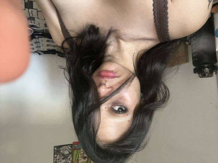 A woman with dark hair and facial piercings poses upside down. She has piercings on her eyebrow, nose, and lip. She's wearing a brown bra with lace trim. A black water bottle is near her head. A blurry object is in the upper left corner. There is a patterned blanket or sheet on the upper left. An art piece is partially visible in the bottom left.
