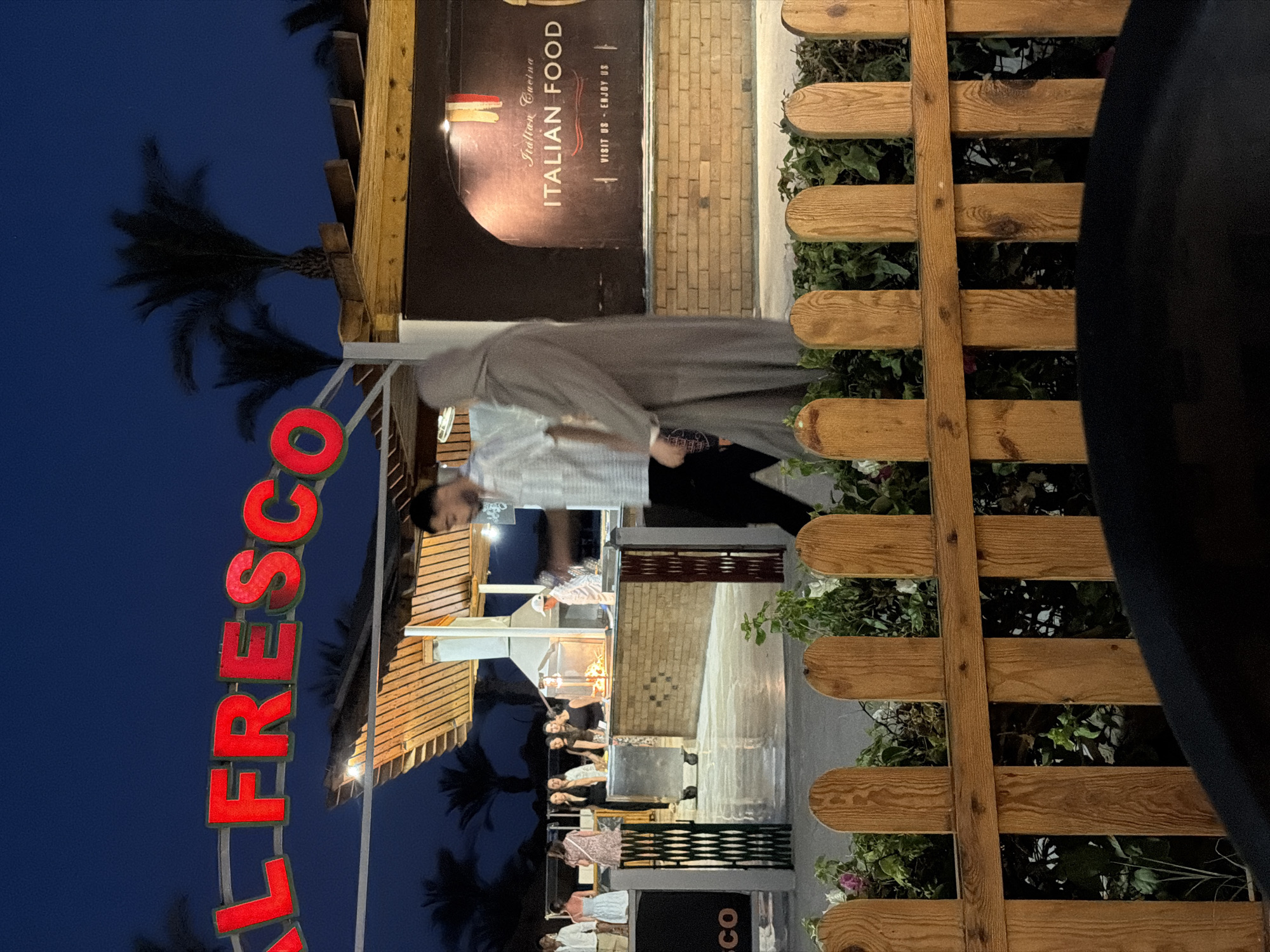 The image depicts an outdoor dining area at night. A large, illuminated red sign that reads "AL FRESCO" is prominently displayed overhead. Beneath the sign, a covered seating area with a thatched roof is visible, where several people are gathered, some appearing to be staff and others patrons. In the background, a restaurant facade features a sign that says "Italian Cucina ITALIAN FOOD VISIT US - ENJOY US." The foreground is partially obscured by a low wooden fence and lush green foliage. Palm trees are silhouetted against the dark blue night sky. The overall scene suggests a lively evening at a restaurant in Sharm El Sheikh, Egypt, likely along the coast, given the presence of palm trees and the outdoor setting.