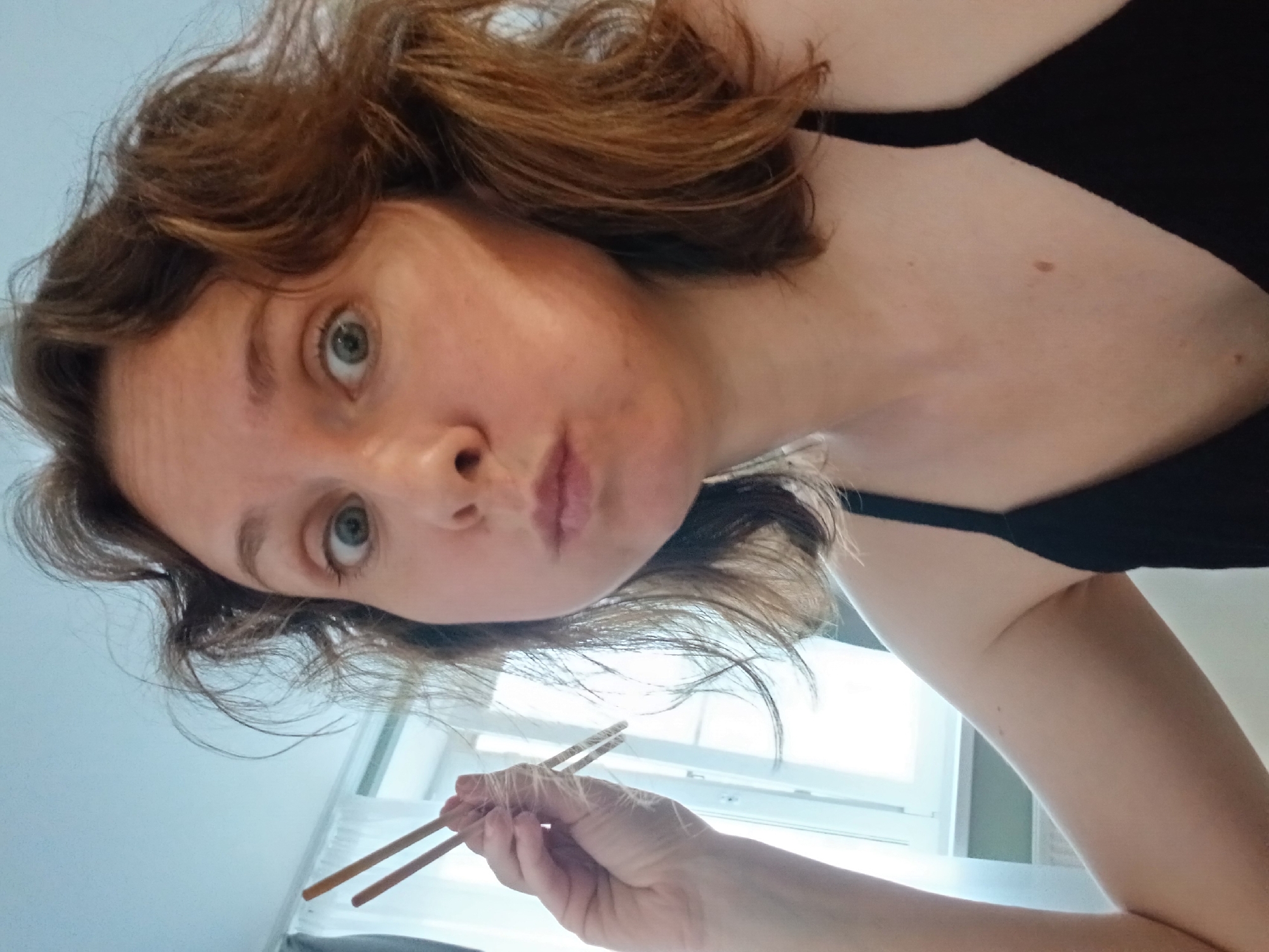 A woman with wavy brown hair and light skin is shown in a close-up selfie. She has wide, surprised eyes and her cheeks are puffed out. She is holding a pair of chopsticks in her right hand, bringing them near her mouth. She is wearing a black spaghetti-strap top. The background is out of focus, showing a window with white curtains and a radiator. There are no discernible landmarks or contextual clues to place the image in Yekaterinburg, Russia.