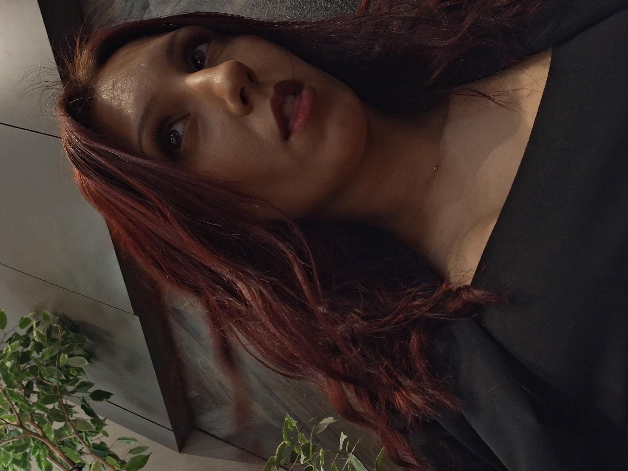 The image presents a head and upper torso shot of a female individual captured from a low-angle perspective in an indoor setting. The subject has light brown skin, dark eyes, and long, wavy reddish-brown hair, with some strands appearing disheveled or windblown. Her gaze is directed upwards and towards the right side of the frame, and her lips are slightly parted, showing a reddish tint. She is wearing a black garment, possibly a top or jacket, which reveals her upper chest and collarbone, where a delicate, thin necklace with a single small bead is visible. The background features dark, paneled walls or surfaces with discernible seams, indicating a modern interior. Green foliage from an indoor plant with small, light-green leaves is visible in the upper left and mid-left areas of the frame. The scene is dimly lit. The location is an indoor establishment in Varna, Bulgaria; however, the precise venue within the city cannot be identified from the visual information provided.