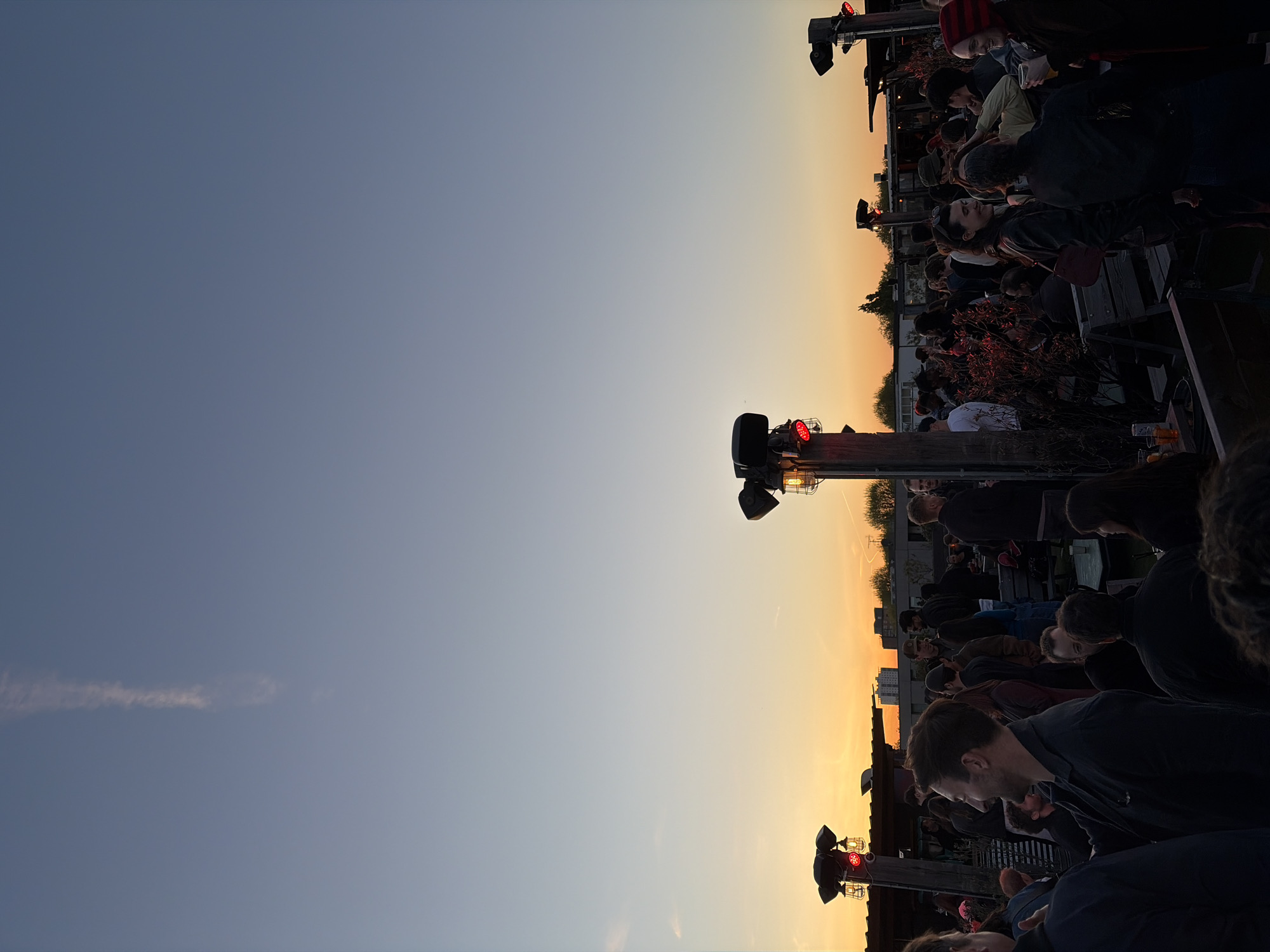 The image depicts an outdoor social gathering at sunset or dusk, likely on a rooftop or elevated terrace in an urban setting. The sky, which occupies the upper half of the frame, transitions from a deep, cool blue-grey at the zenith to warm, vibrant oranges and yellows near the horizon, indicative of late evening. A faint, wispy cloud is visible in the bluer part of the sky.
The lower and right portions of the image are densely populated with a crowd of people, mostly adults, engaged in conversation and interaction. They are casually dressed and appear relaxed, with some individuals holding drinks. Their faces are varied, some in profile, some looking towards the horizon, and others interacting with those around them.
The scene is illuminated by the fading natural light and several artificial light fixtures. These industrial-style lights are mounted on dark poles or structures, featuring bright red warning or accent lights and amber-glowing caged bulbs. Dark, shrub-like vegetation is interspersed among the crowd, possibly serving as decorative elements or low barriers. In the distance, silhouetted against the colorful horizon, are the outlines of buildings, confirming the urban environment. No specific architectural landmarks are discernible, but the overall impression is that of a bustling city at nightfall. No visible text is present.