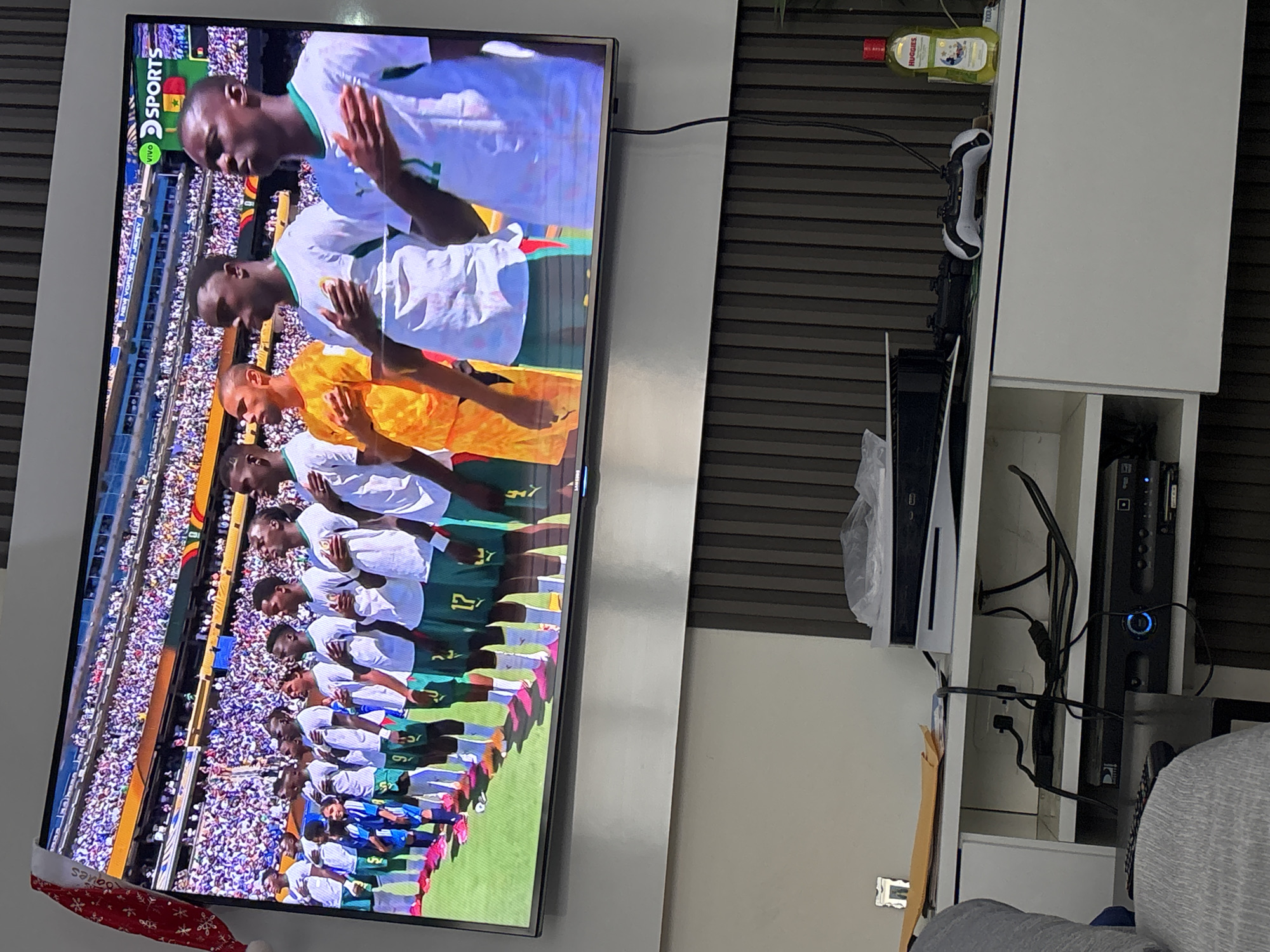 The image shows a television screen displaying a soccer match. The players are lined up on a field, and a crowd is visible in the stadium seating. The team on screen appears to be wearing white and green uniforms. The television is mounted on a wall, and a Christmas hat is draped over the top left corner of the screen. Below the television, a white entertainment unit is visible, holding various electronic devices and accessories, including game controllers and a bottle of Huggies hand sanitizer. The scene appears to be indoors, likely a living room.