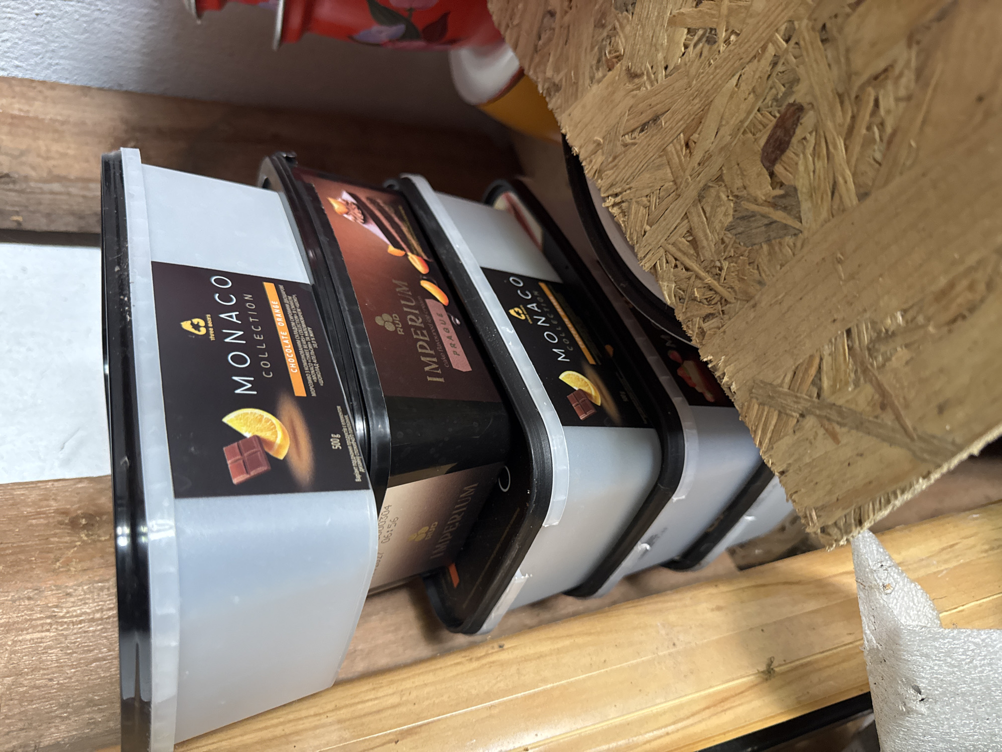 The image shows a close-up view of several tubs of ice cream stacked on a wooden surface, possibly a shelf or pallet. The tubs are mostly covered by their plastic lids, with the labels visible. The dominant visible text on the labels is "MONACO COLLECTION" and "IMPERIUM". One label clearly indicates "CHOCOLATE ORANGE" ice cream with a depiction of chocolate and orange slices. Another label, partially obscured, shows "cake flavoured ice cream PRAGUE". The tubs are predominantly black and clear plastic. The background consists of rough-hewn wood, suggesting a storage area or workshop. There are no people visible in the image, and the lighting suggests it might be indoors with artificial light or a cloudy day. The scene is static, with the ice cream tubs simply resting on the wooden surface. The overall impression is of a retail or storage environment for food products. The text "Rozdilna, Ukraine" provides a specific geographical context.
