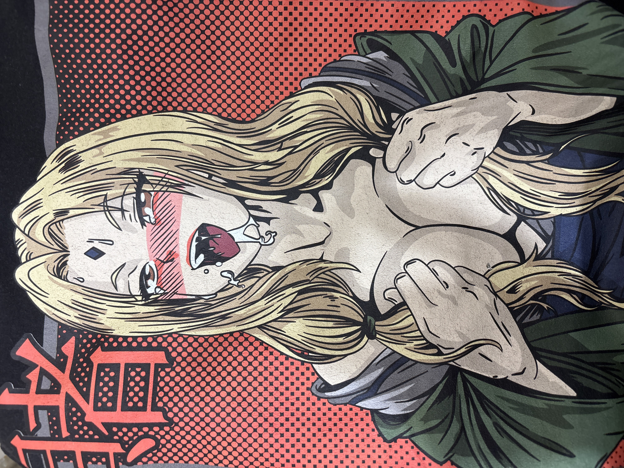 The image shows an anime-style illustration of a blonde woman with exaggerated features such as large eyes, blushing cheeks, and drool. She is holding her breasts with both hands. She is wearing a green garment with grey and blue accents. The background consists of a red dotted pattern. Japanese characters are printed in red and black in the lower left corner. The image is a print on fabric. No information indicates this is in Sofia, Bulgaria.