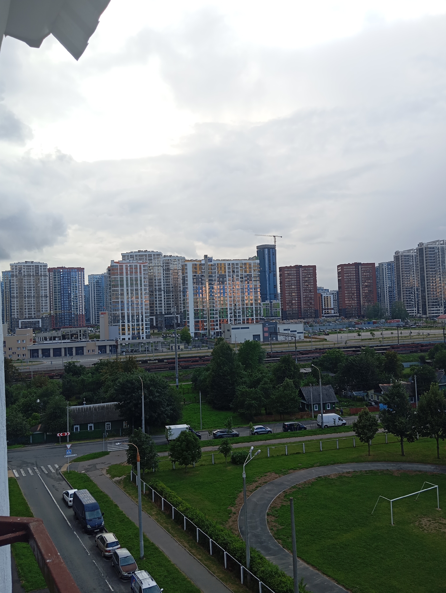 The image is an outdoor, high-angle, daylight shot of a city skyline and surrounding area. In the background, a cluster of modern, multi-story residential buildings dominates the view, some featuring colorful facades. A construction crane is visible behind some of the buildings. In the midground, there are train tracks and several low-rise industrial or commercial buildings. A road runs through the foreground, with several cars parked along its side, including a blue van and several passenger vehicles. Adjacent to the road is a grassy area with a running track and a soccer goal. Trees and vegetation are present throughout the scene, with a few older, smaller houses visible further in the distance. The sky is overcast with visible clouds. The overall impression is a developing urban or suburban area.