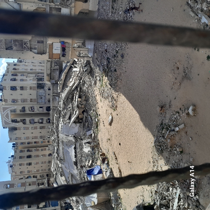 The image depicts a devastated urban landscape in Khan Yunis, Palestinian Territory, viewed through vertical metal bars, likely from a window or balcony.
In the foreground, two out-of-focus, twisted vertical metal bars frame the scene. The ground immediately visible is predominantly sandy earth with scattered concrete rubble and debris. A prominent shadow extends diagonally from the right across the sandy area.
The midground is dominated by massive piles of concrete rubble, twisted rebar, and debris, indicative of collapsed multi-story structures. The remnants of a large, possibly domed or curved concrete structure are discernible amidst the debris. To the far left, a white tent structure is visible, adjacent to which lies a crumpled blue tarp or fabric.
In the background, several multi-story apartment buildings stand, all exhibiting extensive damage. Facades show numerous impact points, missing sections, blown-out windows, and exposed interiors. Many windows are shattered or missing. One building on the far left features a distinctive light-colored arched architectural element at its roofline. On a damaged balcony of a building to the right, a line of laundry is hanging.
In the lower right quadrant of the image, a single individual, wearing dark clothing, is captured in motion, running from right to left across the sandy ground. The sky above is partially cloudy, indicating daylight conditions. A "Galaxy A14" watermark is present in the lower left corner of the image.