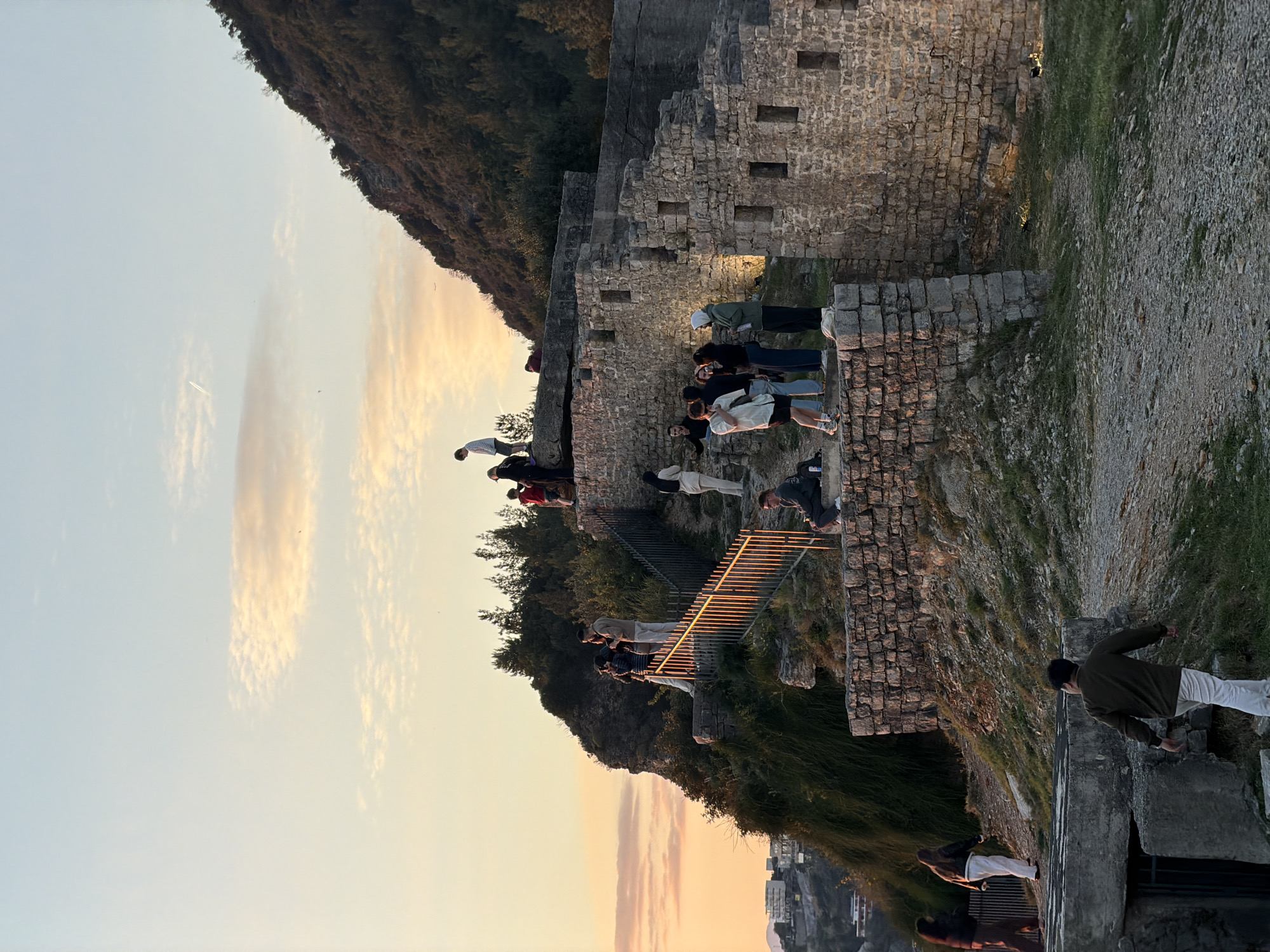 The image shows a view from the Citadel of Budva, Montenegro, during what appears to be sunset. The sky is a mix of light blue and orange hues.
A group of people are gathered along the stone walls and a metal staircase. They seem to be observing the view. The stone structure suggests a historical fortification.
On the left, a hill covered in vegetation is visible. Buildings are seen in the distance. The light and shadow indicate the time of day.