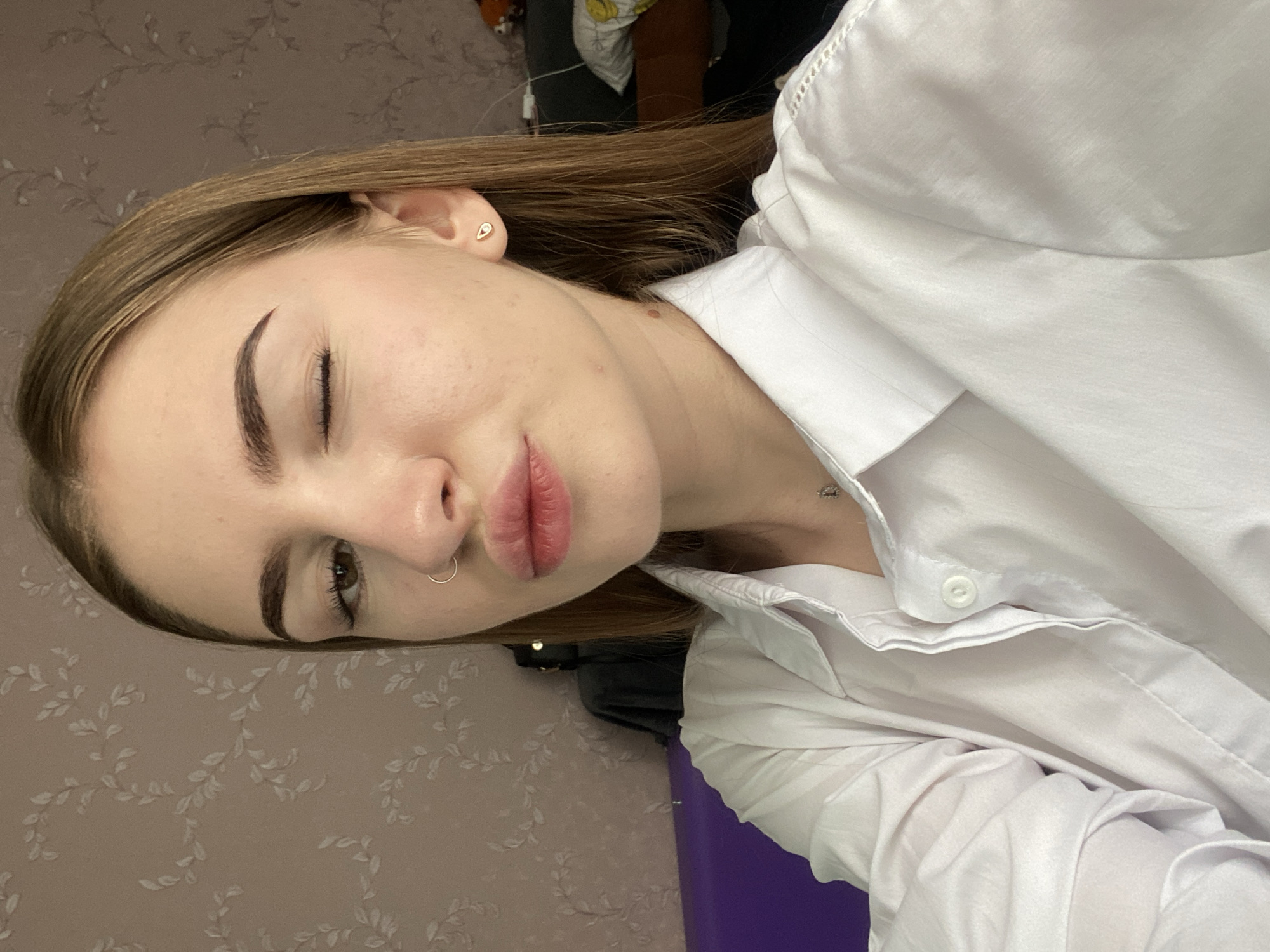 A young woman with long, light brown hair is shown in a close-up selfie. She is wearing a white collared shirt and has a nose ring. Her lips are pursed as if she is about to kiss, and she is winking with her right eye. A small teardrop-shaped earring is visible on her left ear. The background appears to be a room with patterned wallpaper in shades of pink and beige. To the left, a section of a purple piece of furniture is visible. The scene is indoors and does not provide specific geographical markers for Дьяково, Russia.