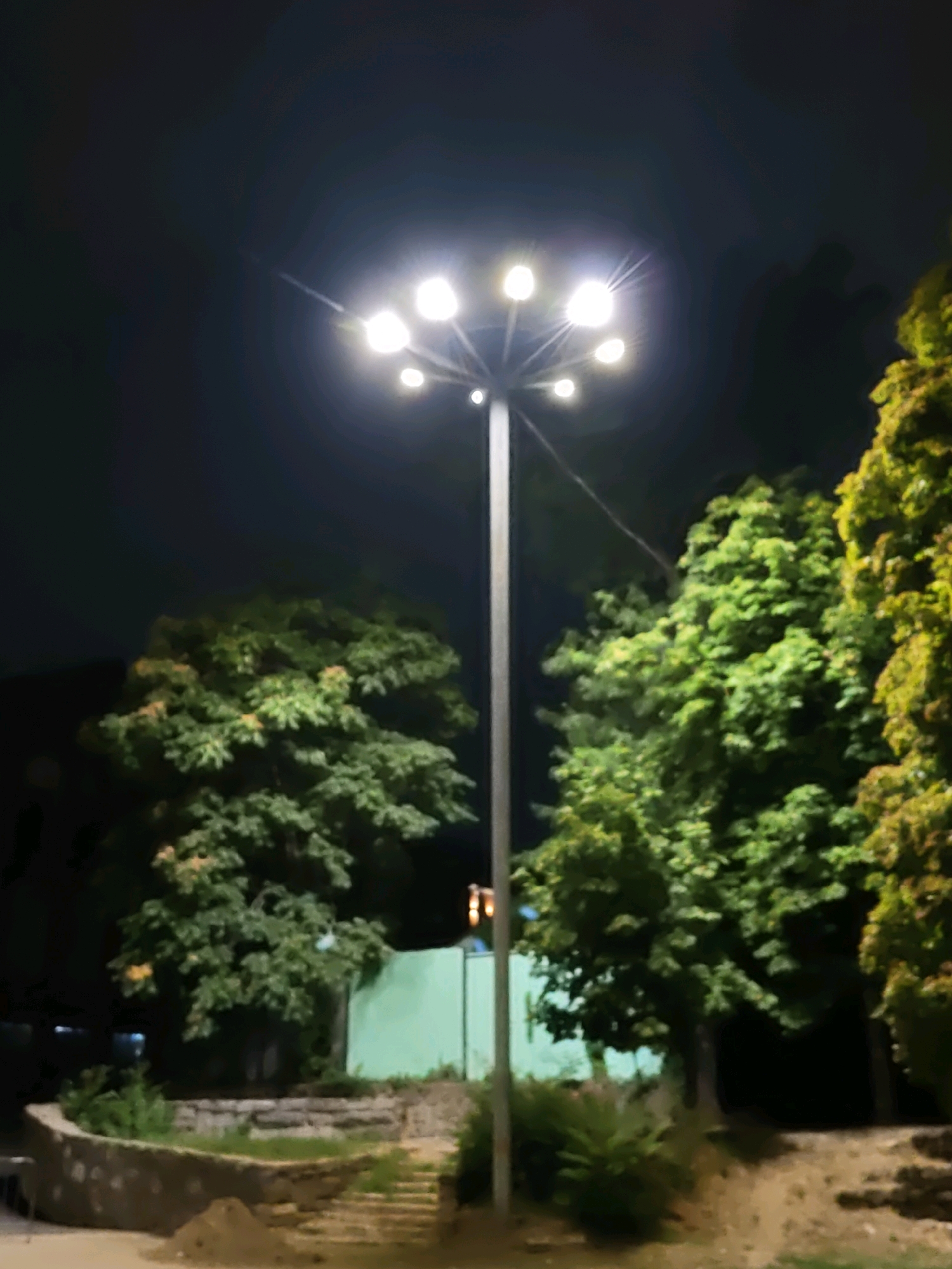 This image depicts an outdoor scene at night, illuminated by a tall street lamp with multiple lights at its apex. The lamp is positioned centrally in the frame and emits a bright, diffused light that creates a halo effect. Surrounding the lamp are several large, leafy trees with dense green foliage. To the left of the lamp, there is a curved, low stone wall with some vegetation growing behind it, and a set of stone steps leading upwards. In the background, a light green fence is visible. The ground in the foreground appears to be unpaved, with some dirt or sand. The overall impression is of a park or public area at night. There are no people visible in the image. Based on the provided context, this scene is located in the city of Rostov-on-Don, Russia.