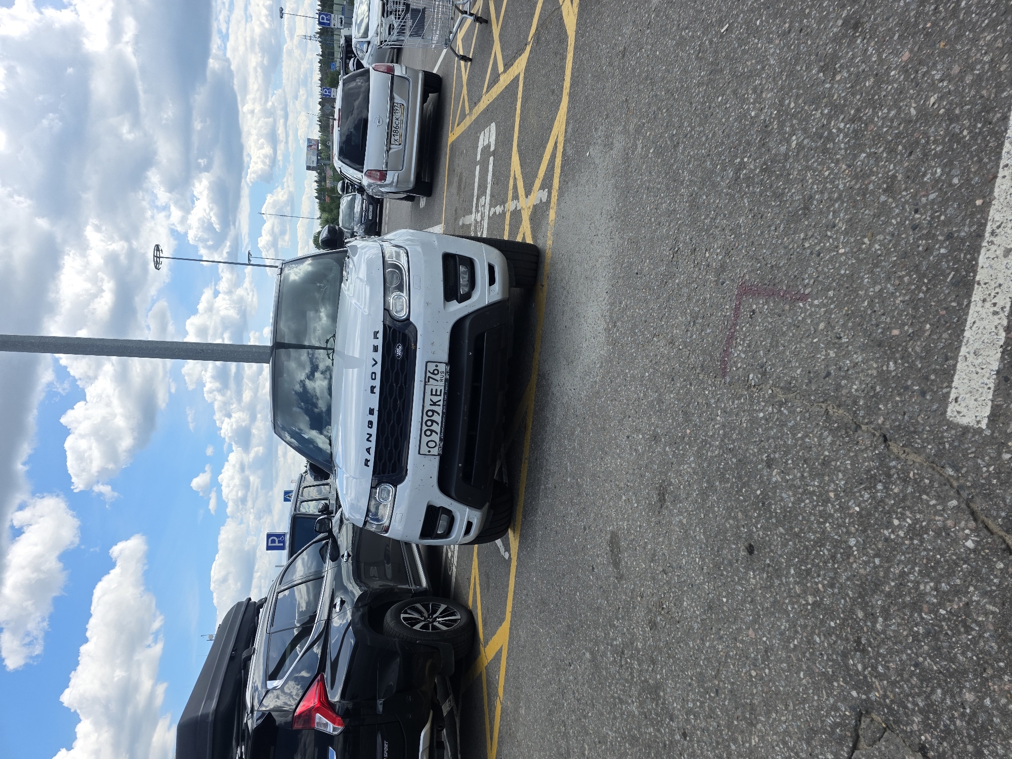 This is a vertical, outdoor, daytime photograph taken in Sholokhovo, Russia. The scene depicts a parking lot with several cars parked in designated spots marked with yellow lines. The foreground is dominated by a white Range Rover, its front end angled towards the viewer. Behind it, a black SUV with a roof box is parked. Further back, other vehicles are visible, including a silver car with a license plate that reads "X186CK 197". The sky is filled with fluffy white clouds against a bright blue backdrop, suggesting fair weather. Tall light poles are scattered throughout the parking lot. The ground is asphalt. Text visible includes the word "ROVER" on the front of the white vehicle, a license plate that reads "O 0999 KE 76 RUS", and a blue parking symbol sign. The overall impression is that of a typical parking scene on a sunny day.