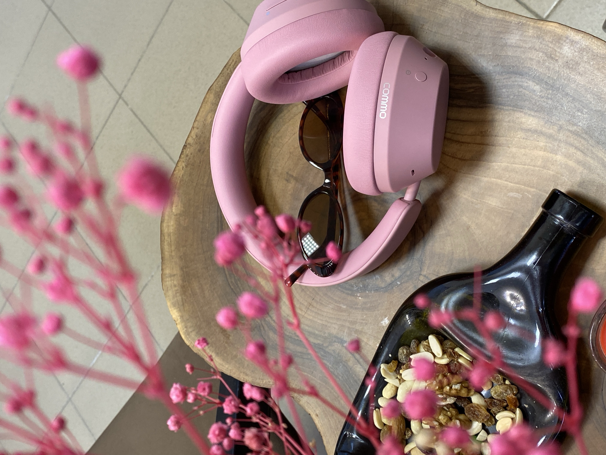The image depicts an overhead shot of a wooden surface, possibly a tabletop, with several items arranged on it. Dominating the composition are a pair of pink Commo headphones and a pair of tortoiseshell sunglasses nestled within the headphones. A black, oddly-shaped dish filled with mixed nuts and raisins sits to the side. A blurred foreground element of pink flowers adds a soft, out-of-focus frame. The lighting suggests an indoor setting, possibly near a window. There are no people visible, and the scene appears staged. No information on the city of Votkinsk, Russia is available.