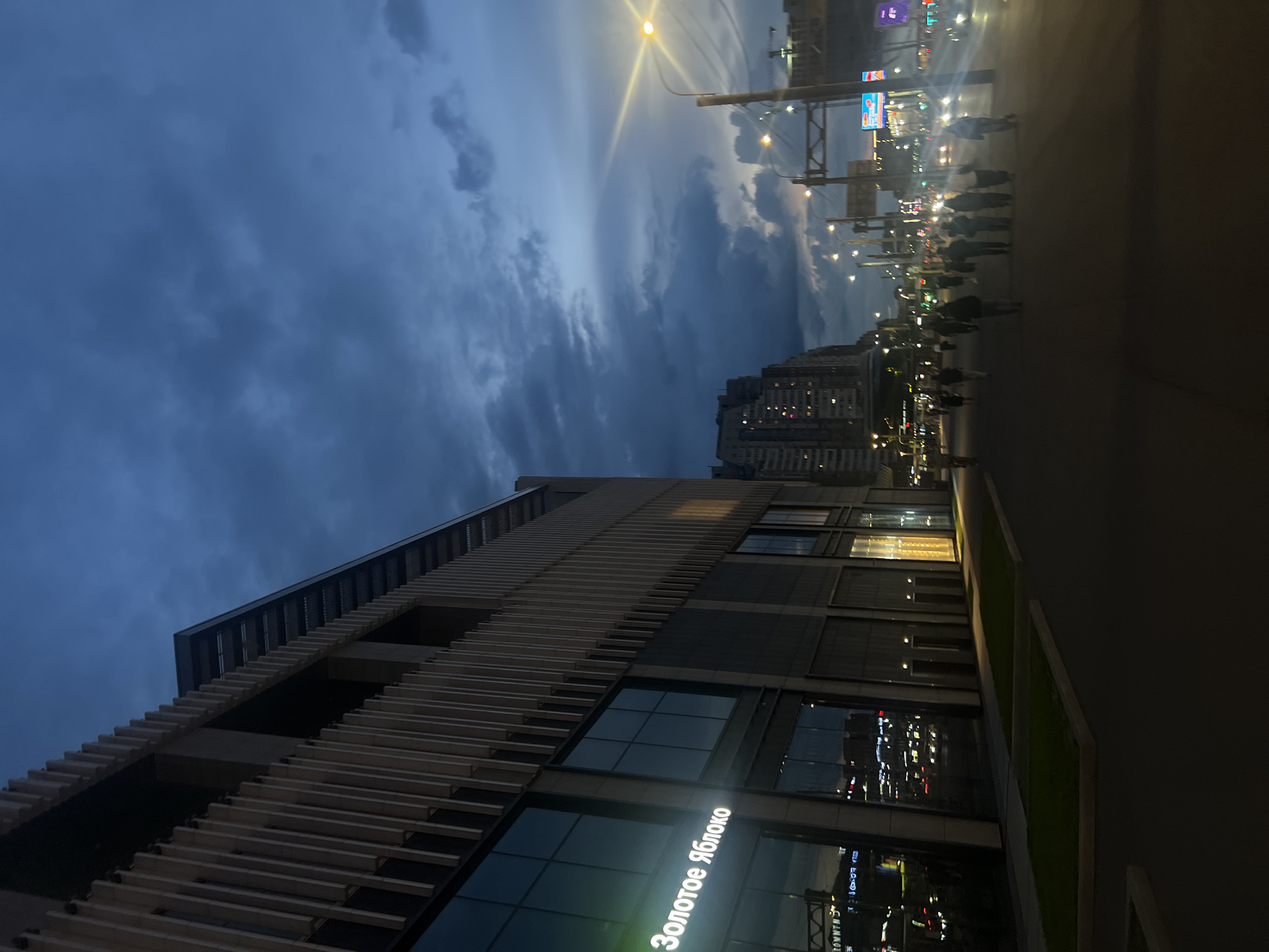 This image captures an urban street scene at dusk or early night in Saint Petersburg, Russia, specifically depicting the Zolotoye Yabloko (Golden Apple) store located on Ligovsky Prospekt 30A.
The left half of the image features a modern multi-story building with a facade characterized by numerous vertical, light-colored slats or fins extending upwards, interspersed with dark, reflective glass window sections. A prominent, illuminated sign in Cyrillic, "Золотое Яблоко" (Golden Apple), is visible on the lower section of this building's glass frontage. Reflections of distant city lights and traffic are discernible on the building's dark windows. At the base of the building, a dark paved pedestrian area is present, featuring several rectangular planter boxes containing patches of dark green grass.
The sky occupies the upper portion of the frame, displaying a deep blue-grey hue with prominent, heavy cloud formations, indicative of twilight or an overcast evening.
In the midground and extending towards the right, a wide street or thoroughfare is visible. Approximately 15 to 20 individuals, mostly seen from behind, walk along the paved surface, generally moving away from the viewer. Their forms are largely silhouetted against the ambient and artificial light, rendering individual features indistinct. Further right, vehicle headlights and taillights appear as bright, elongated streaks, suggesting moving traffic on the road. Urban infrastructure includes multiple streetlights emitting bright yellow-orange light, some with visible overhead cables, and a large, dark metallic gantry structure spanning across the street. Distant buildings with illuminated windows are visible on the far side of the road, receding into the urban landscape under the low light.
