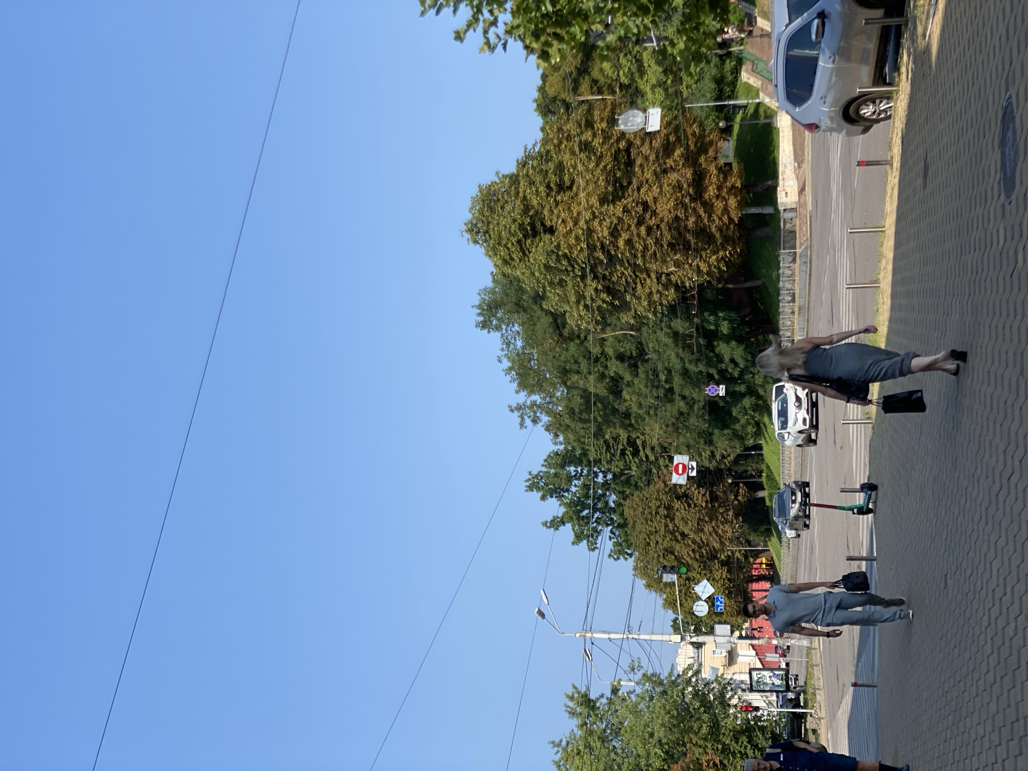 In a city scene under a clear blue sky, a paved sidewalk runs along a street with traffic. Several people are walking on the sidewalk, including a man in a gray polo shirt and pants carrying two bags, and a woman in a gray dress holding a black bag. Further down the sidewalk, another person is visible. On the street, cars are driving and stopped at an intersection with traffic lights and signs. An electric scooter is parked on the sidewalk. Trees line the street, with some showing autumnal colors. Buildings and infrastructure, including streetlights and power lines, are visible in the background.
