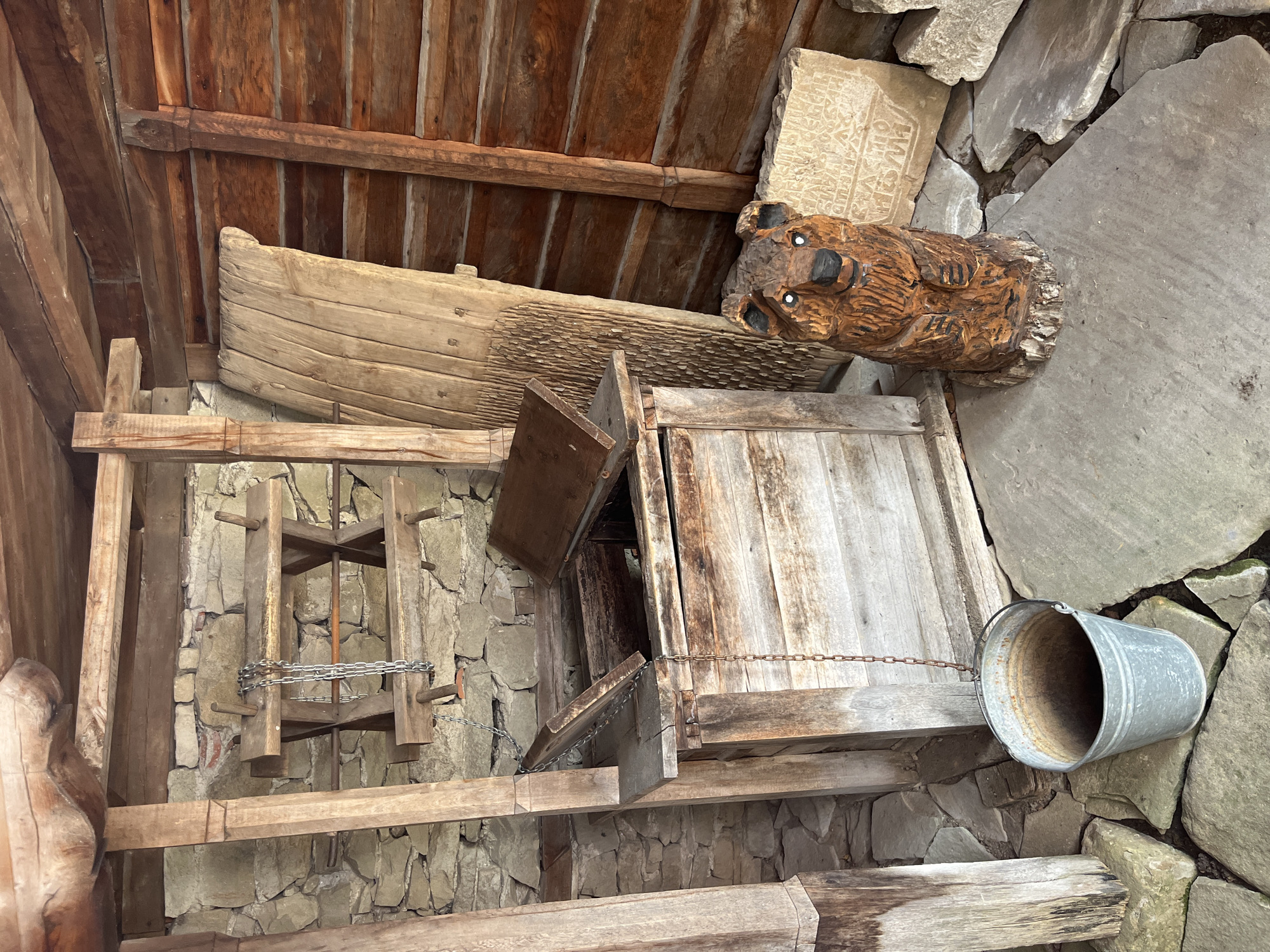 In Zheravna, Bulgaria, an old well is housed within a rustic wooden structure. The well itself is made of weathered wood, with a hinged lid partially open, revealing a dark interior. A metal bucket hangs from a chain attached to the well's structure, positioned on the stone-tiled ground below. To the right of the well stands a carved wooden bear, its features detailed with painted eyes and a black nose. Behind the bear, a carved stone slab with inscriptions leans against the wooden wall of the structure. The walls of the enclosure are constructed from rough stone and rough-hewn timber logs. Various wooden planks and tools are visible within the structure, suggesting it might be a preserved historical exhibit or a functional space from a bygone era. The scene is lit by natural light, casting shadows and highlighting the textures of the aged materials.