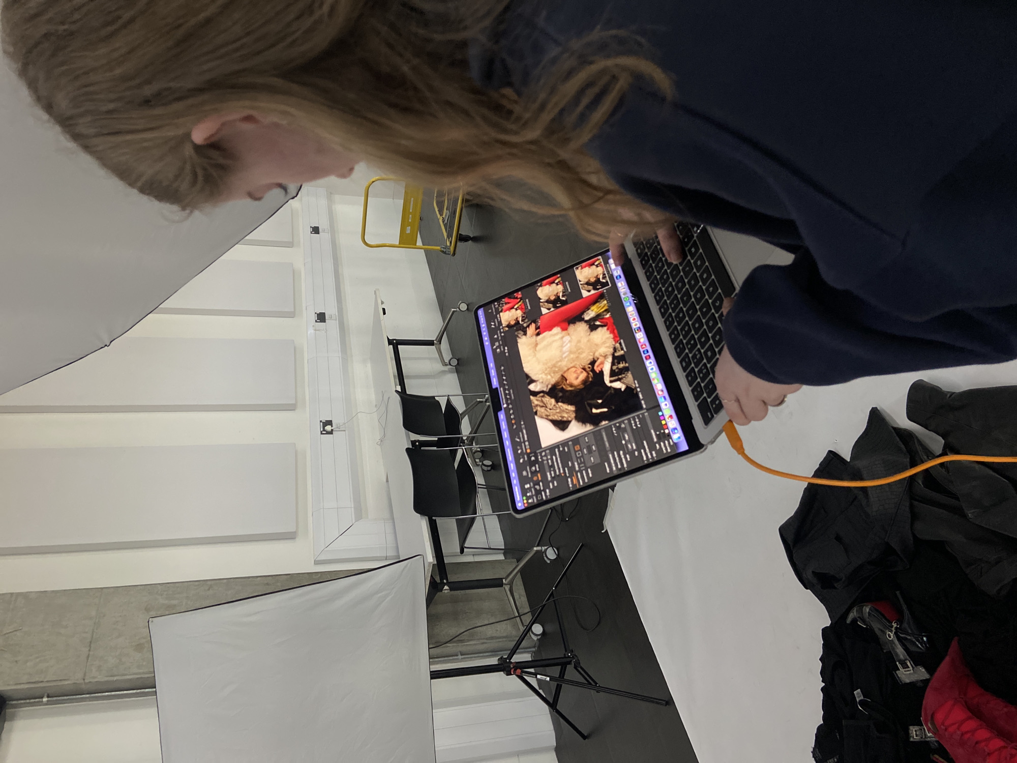 A blonde-haired individual wearing a dark blue sweatshirt is observed from a high angle, focusing on a laptop screen. The screen displays images of a person in a white fuzzy coat with red boots. The individual's hands are on the laptop keyboard. An orange cable connects to the laptop. The environment is a photo studio with white diffusion panels, black chairs, and clothing items on the floor. There are white rectangular panels on the wall. A yellow handcart is visible in the background. The location appears to be indoors. There is no way to verify if this is London, UK.