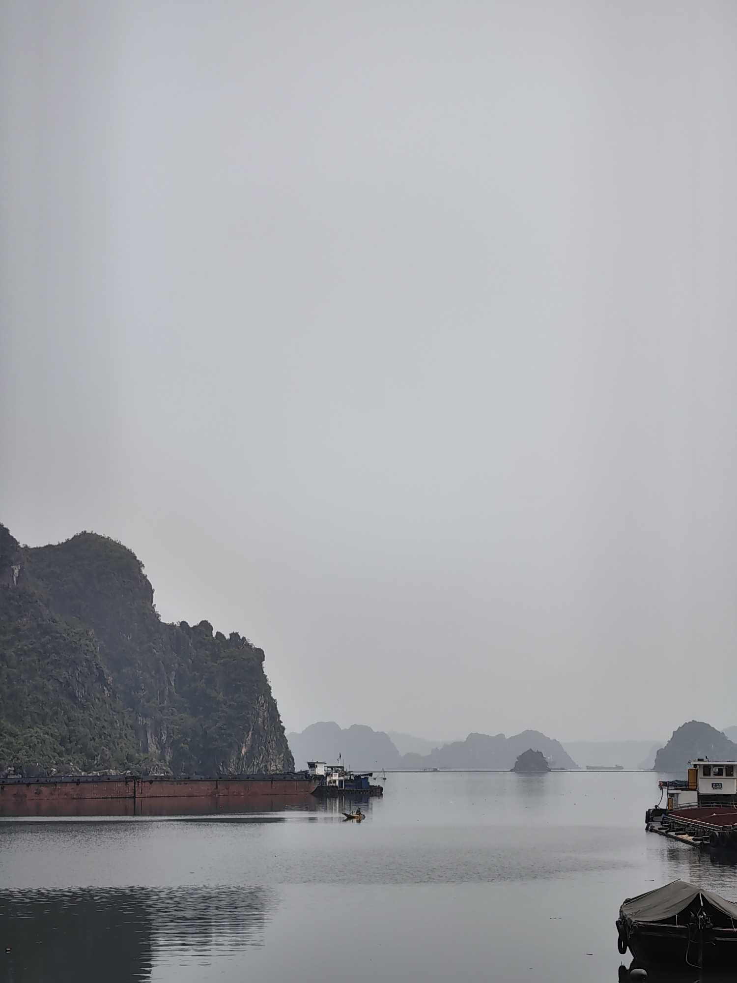 The image depicts a hazy, overcast day on the water near Quang Hanh Ward, Vietnam. Several islands and a large, green, mountainous land mass are visible in the background. In the foreground, there are multiple boats and barges. One large, reddish-brown barge is positioned near the mountainous land mass. Further out, another boat is present with a smaller boat near it, and to the right is a boat docked with a covered vessel in the lower right corner. The water is calm with reflections of the sky and surrounding objects.