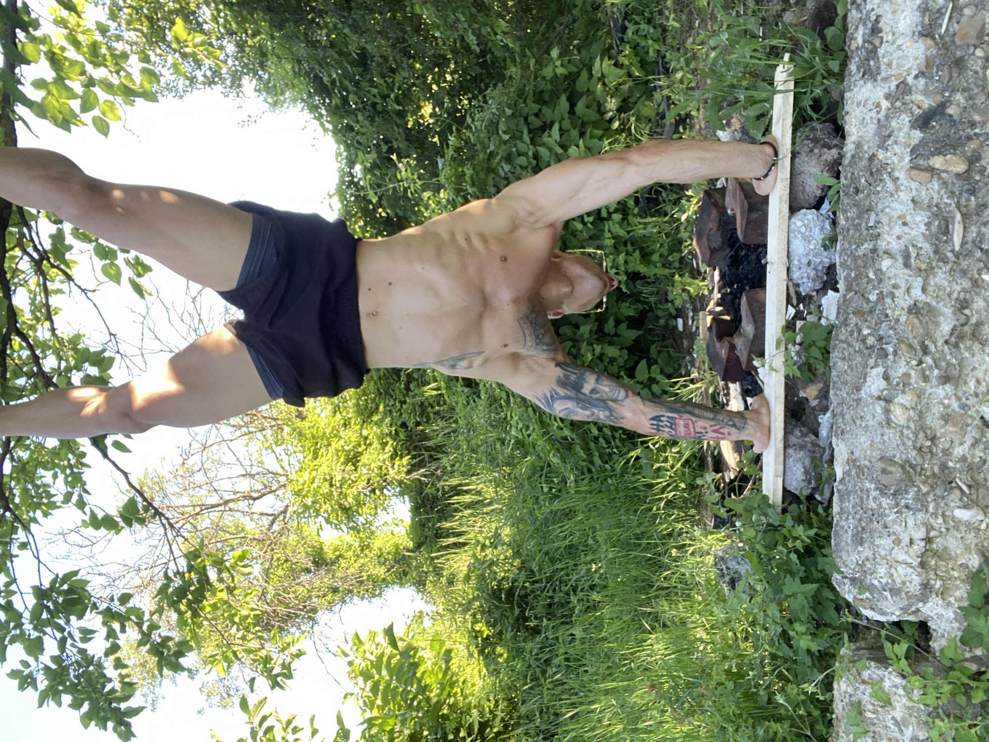 A muscular man with tattoos on his arms and legs performs a handstand outdoors, supported by his hands on a thin wooden plank. He is positioned with his feet in the air, his legs extended upwards, and his head pointed downwards. He wears black shorts. The background consists of lush green foliage, trees, and a bright sky, suggesting a natural, possibly park-like setting. A small fire pit with bricks is visible behind him. The foreground features rough, textured concrete blocks.