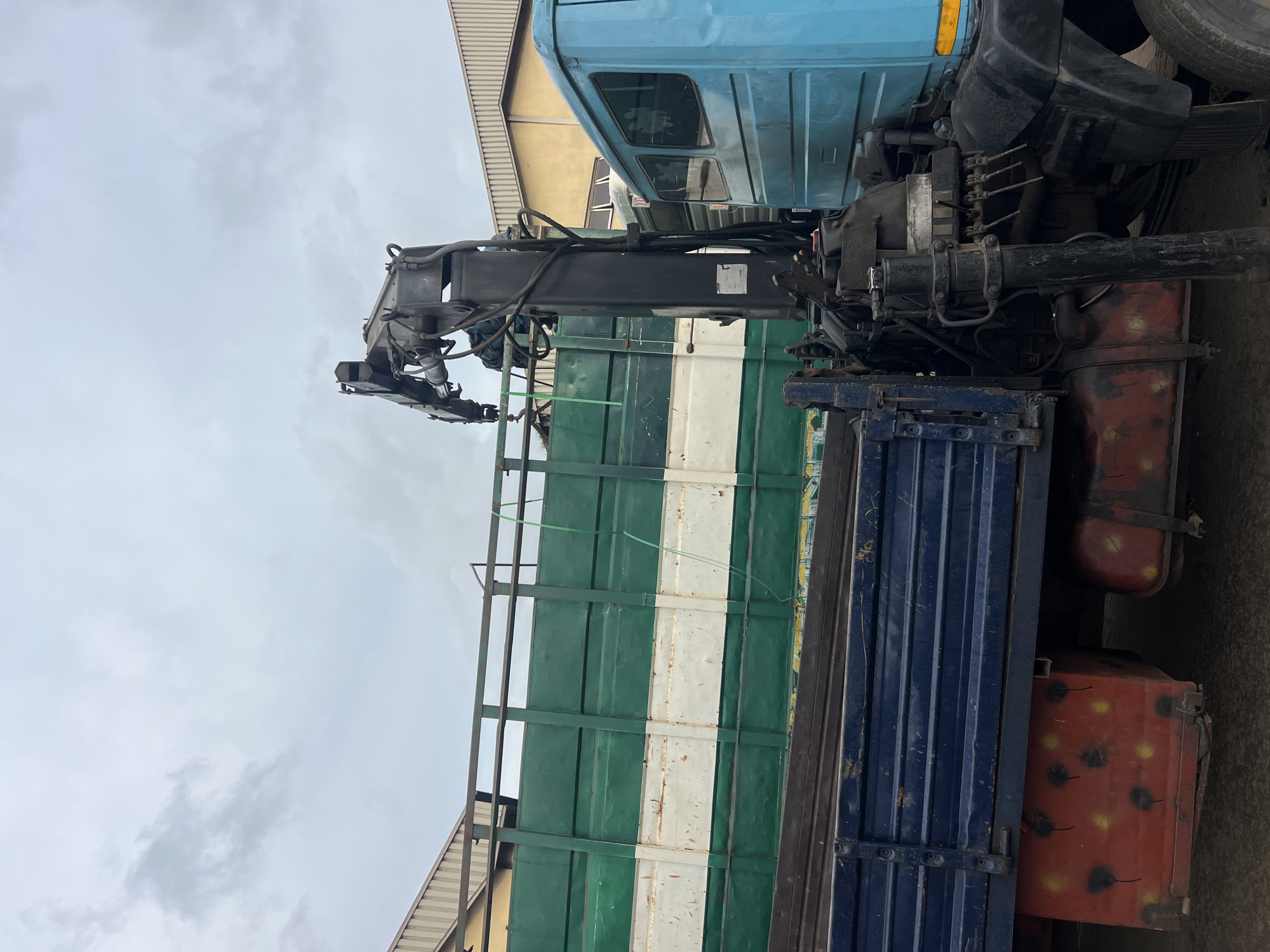 The image shows a truck with a crane mounted on the back. The truck bed is divided into sections, with green, white, and blue sections. The fuel tanks are red and orange. The sky is overcast. The photo likely taken in the city of Lagos, Nigeria based on the image quality and the truck type commonly found there.