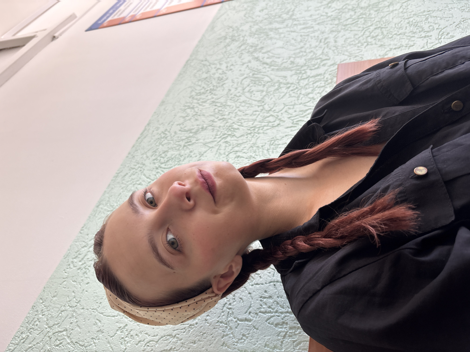 A young woman with reddish-brown hair in two braids, wearing a black shirt and a beige headscarf with polka dots, is looking at the camera. She is seated in an indoor environment with a textured light green wall behind her. The background also shows a white ceiling and a hint of a poster on the right. The lighting suggests it is daytime. The image appears to be a selfie taken by the woman. The location is not identifiable as Chaikovsky, Russia, from the visual information provided.