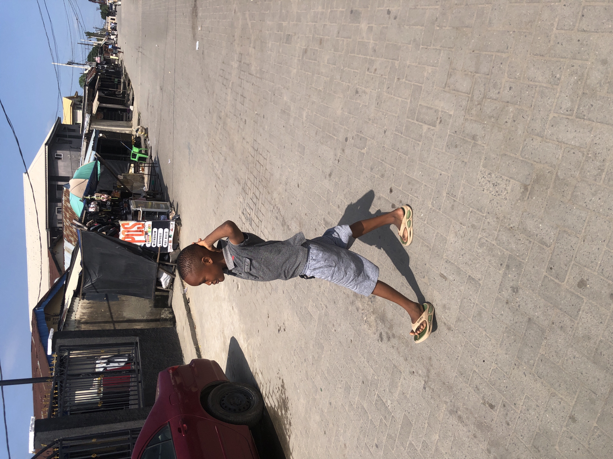A young boy walks across a paved street in Ado, Nigeria. He is wearing a gray polo shirt and gray shorts, with light-colored flip-flops on his feet. The boy is facing to the right of the frame, with his left arm behind his head. The street is lined with small shops and buildings. A red car is parked on the left side of the street. The sky is clear and blue.