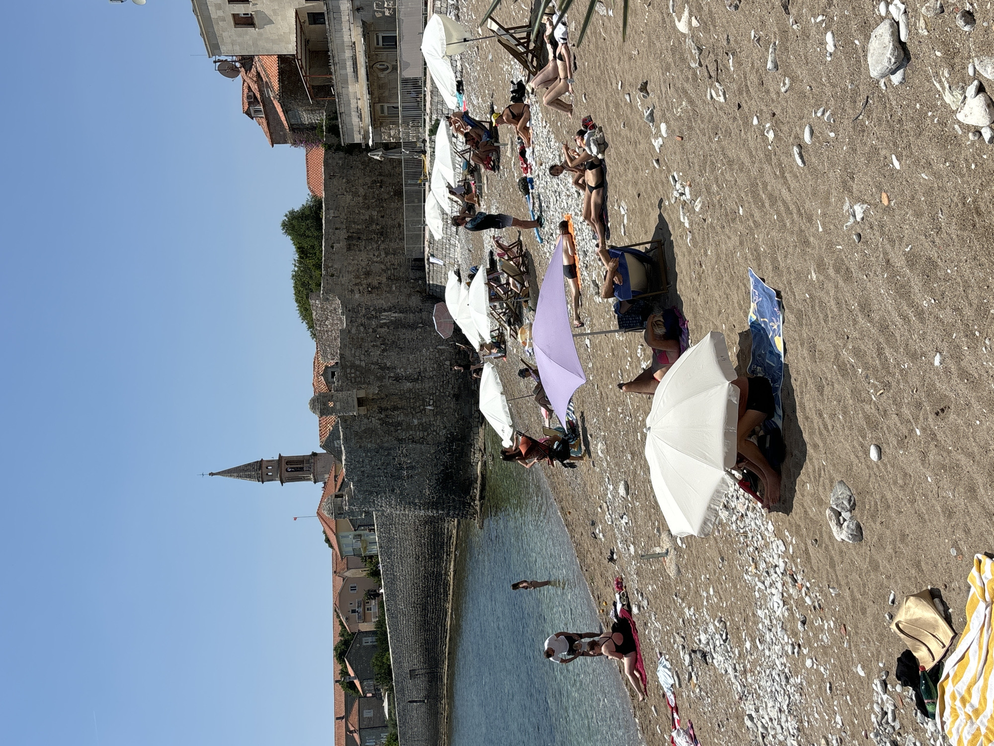 This is a tilted photograph of a crowded beach on a sunny day in Budva, Montenegro. The beach is composed of sand and small white pebbles, with a clear, calm body of water to the left. Several people are scattered across the beach, sunbathing on towels and chairs, and relaxing under umbrellas. Some individuals are in the water, wading or swimming. The backdrop features old stone fortifications and buildings with red tile roofs, suggesting a historic coastal town. A church steeple is visible in the distance, further identifying the location. The bright sunlight and clear blue sky indicate it is daytime. The overall mood appears relaxed and leisurely, as people enjoy a typical summer beach day. There is no visible text in the image.