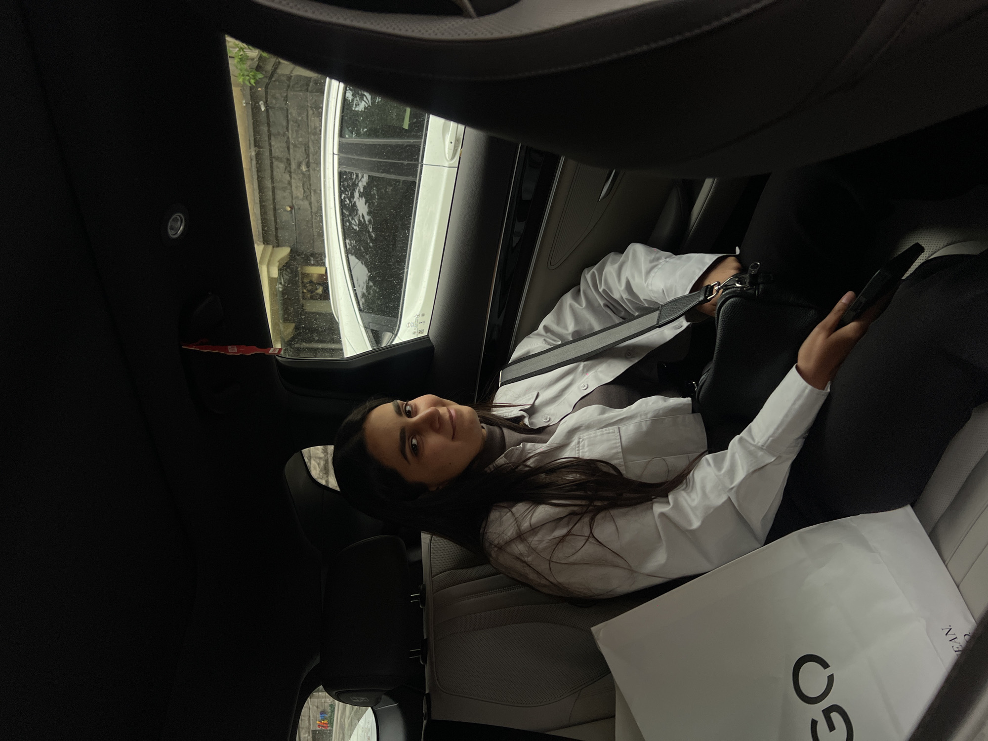 A woman is lying down in the back seat of a car. She has dark hair and is wearing a white shirt, a seatbelt, and dark pants. She is holding a black purse in one hand and a phone in the other. A large white shopping bag with the letters "GO" printed on it is next to her. The car appears to be parked next to a building with a white car parked along the curb. The location of the scene is in Yerevan, Armenia.