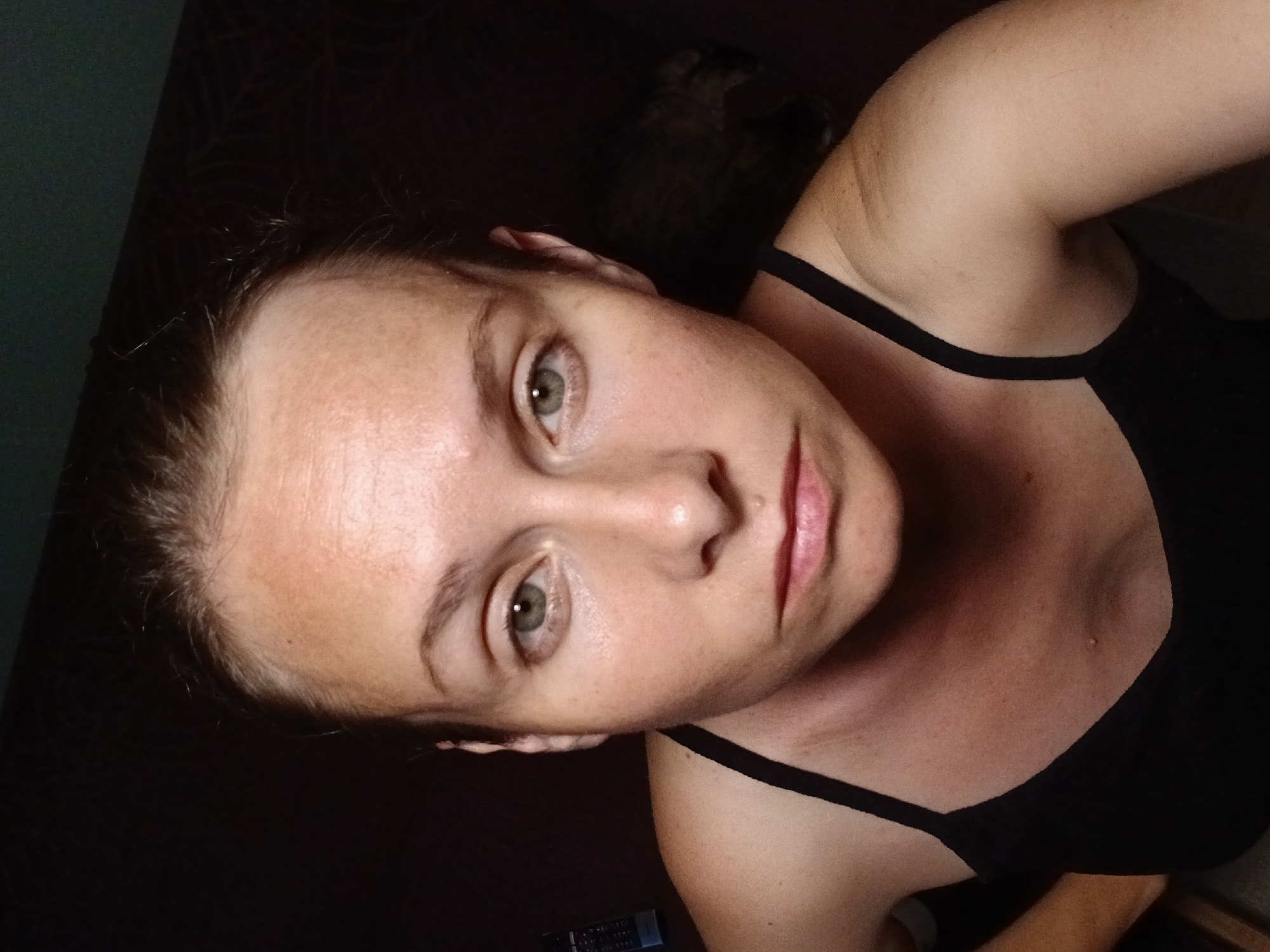 The image shows a medium shot of a Caucasian woman with light brown hair pulled back. She is wearing a black tank top. Her skin appears fair with some freckling on her forehead and cheeks. Her eyes are green, and she has a neutral expression. The background is dark and appears to be indoors, with a hint of a dark object on the right side of the frame, possibly an animal or furniture. A remote control is visible on the left side of the frame. There is no information available about the specific location in Yekaterinburg, Russia, or any actions or interactions occurring in the image.