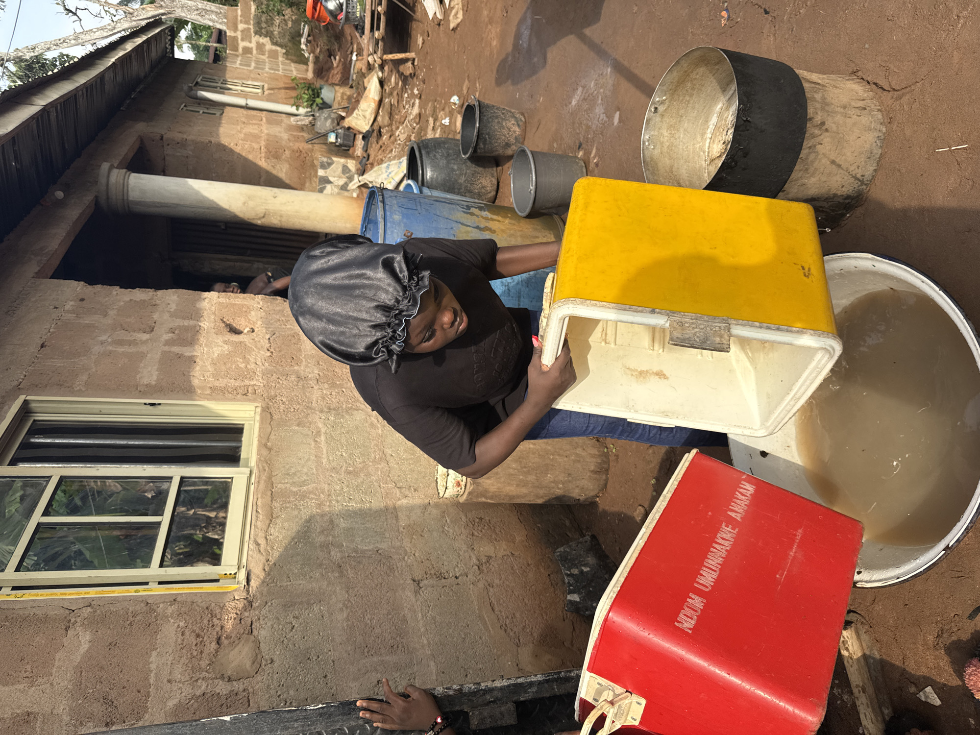 A woman wearing a black head covering is seen holding a yellow and white cooler, with a red cooler bearing the name "NDOM UMLINWAKKE AMAKAM" nearby. Various containers including buckets and drums surround her. She appears to be outdoors, adjacent to a building with a window. The location is Eziudo, Nigeria.