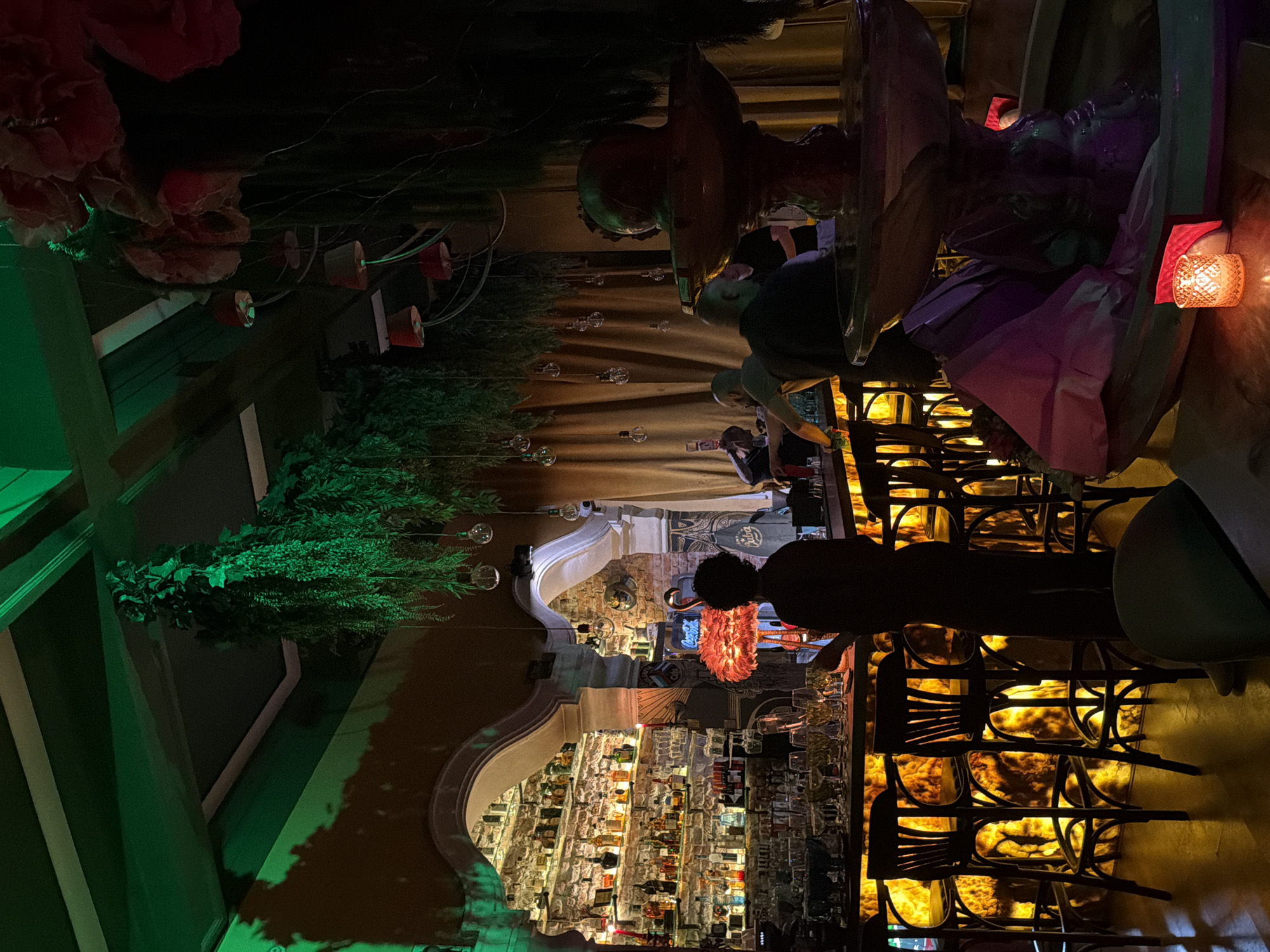 The image depicts the interior of a bar in Sofia, Bulgaria. There are numerous liquor bottles displayed on shelves behind the bar, which has a decorative arch above. Patrons are seated on dark-colored chairs at the bar. The bar top has a glowing, amber-colored material, possibly onyx or a similar stone. Ornate decorations including plants, lights, and what appears to be a fountain with decorative elements are visible overhead. People are interacting behind the bar, likely staff preparing drinks. The lighting is dim with accents of red and amber.