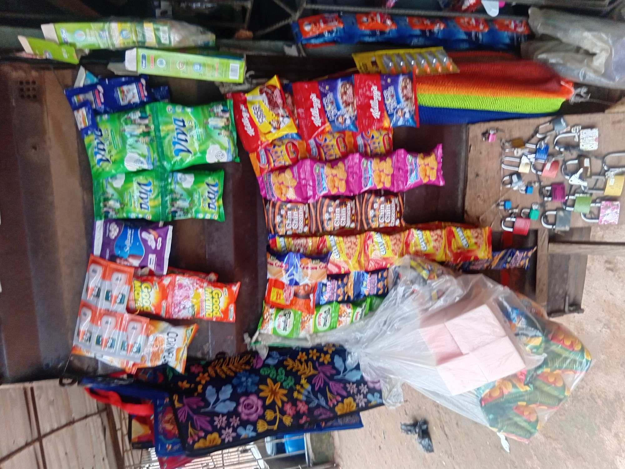 The image depicts a roadside stall in Nkpor, Nigeria, displaying a variety of goods. In the foreground, an assortment of padlocks are laid out on a wooden surface, some with keys. To the left, a large plastic bag contains stacked pink boxes and numerous smaller, colorful packages, likely snacks. The stall itself is constructed from wood, with shelves holding rows of packaged food items and toiletries, including brightly colored bags of "Viva Plus" detergent, packets of "Good Mama" snacks, and various cereal boxes from brands like Kellogg's. A vibrantly colored fabric, resembling a rainbow, hangs on the right side of the stall. The overall setting suggests a typical street market scene in Nigeria.