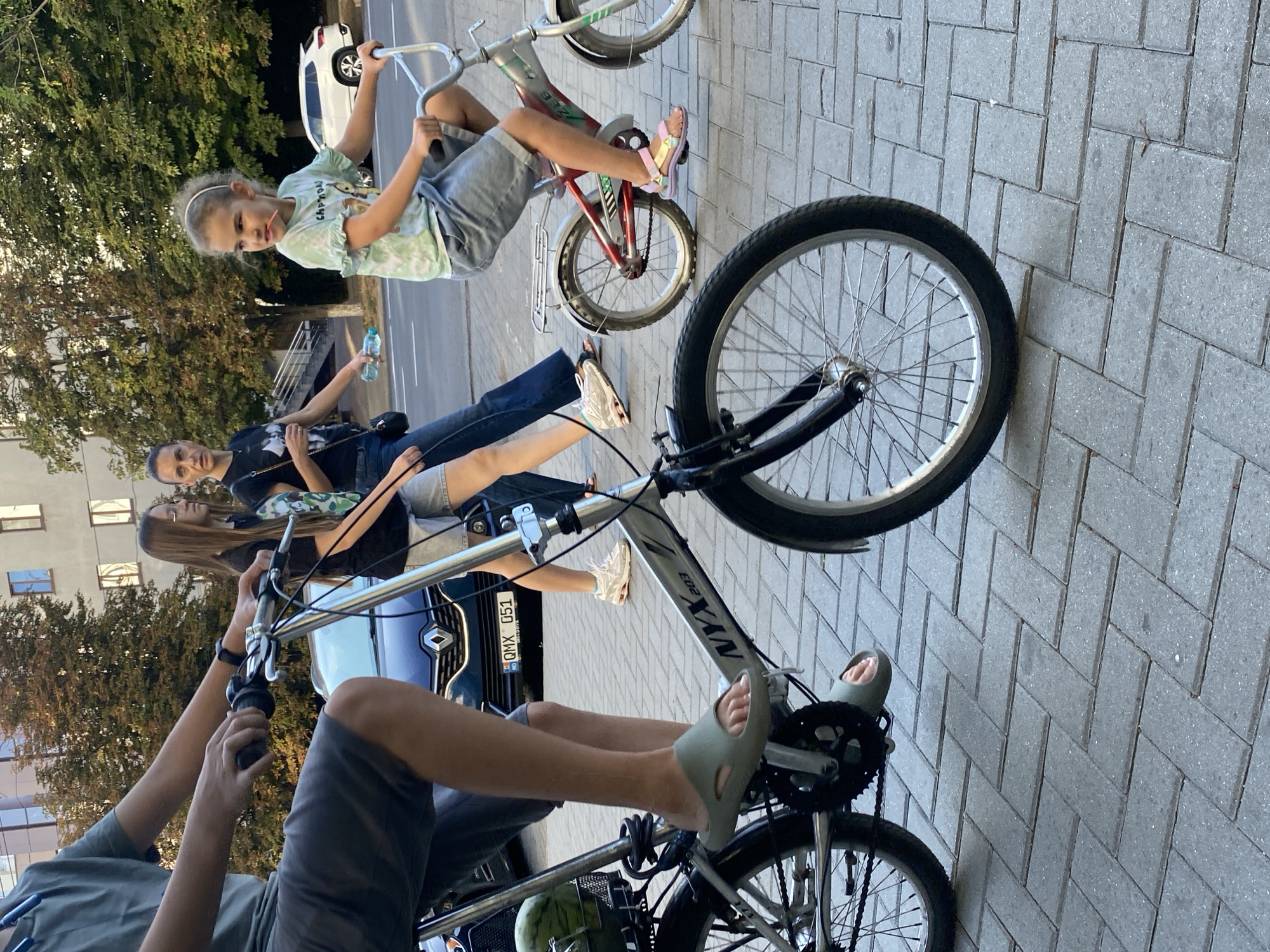 The image shows a street scene in Chișinău, Moldova. In the foreground, a person is riding a silver bicycle with a basket containing a watermelon. The bicycle has the brand name "NYX 203" visible. Further back, a young girl is riding a bicycle, wearing a green and white tie-dye t-shirt and denim shorts. Two women are walking behind her. One of the women is holding a water bottle. In the background, a blue car with a Moldovan license plate (QMX 051) is visible, and beyond that, a white SUV is parked on the side of the road. Buildings and trees line the street, and the ground is paved with grey interlocking bricks. The lighting suggests it is daytime, likely late afternoon due to the long shadows.