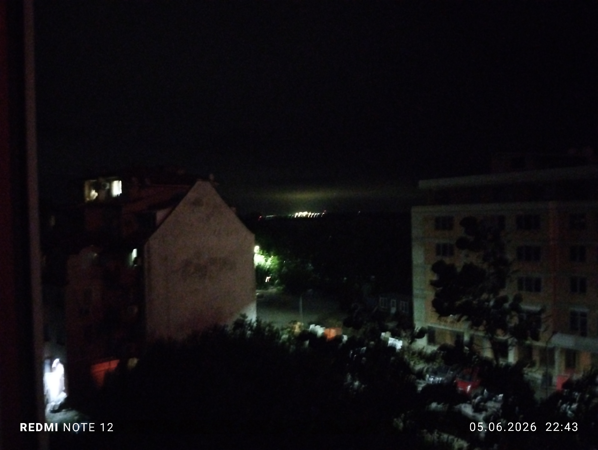 The image depicts a nocturnal urban scene captured from an elevated interior vantage point, likely a window or balcony, on June 5, 2026, at 22:43. The sky is dark, indicating late evening.
In the foreground, a dark vertical element, consistent with a window frame or structural beam, partially obscures the left side. Below it, a bright white light emanates from a ground-level opening or room. Dense, dark vegetation, appearing as trees and bushes, occupies the immediate mid-ground, mostly silhouetted against ambient light.
To the left of the frame's center, a light-colored building with a sloped roof is visible, its facade partially illuminated. Behind this structure, another building shows a single brightly lit window on an upper floor. Towards the right-center, a multi-story residential building with a reddish-brown facade and numerous windows, some faintly illuminated, stands prominently. Directly in front of this building, a dark tree or large shrub obscures parts of its lower sections. Below this building, blurry shapes, including what appears to be a red vehicle, are discernible among the foliage and ground elements. Lighter, indistinct objects are scattered on the ground.
On the distant horizon, a distinct horizontal band of artificial lights is visible, suggesting a distant urban area or a coastline lined with lights. A subtle, diffuse glow is present in the sky directly above this light band.
No individuals are visibly present or engaged in discernible activities. The image quality, as indicated by the "REDMI NOTE 12" watermark, is consistent with mobile phone photography under low-light conditions.
The generic urban architecture and distant lights prevent precise street-level localization within Burgas, Bulgaria. However, the scene is consistent with a view from a residential building within the city, potentially overlooking urban sprawl extending towards the Burgas Bay or one of the city's lakes, where distant lights and a faint horizon glow would be observed from elevated positions.