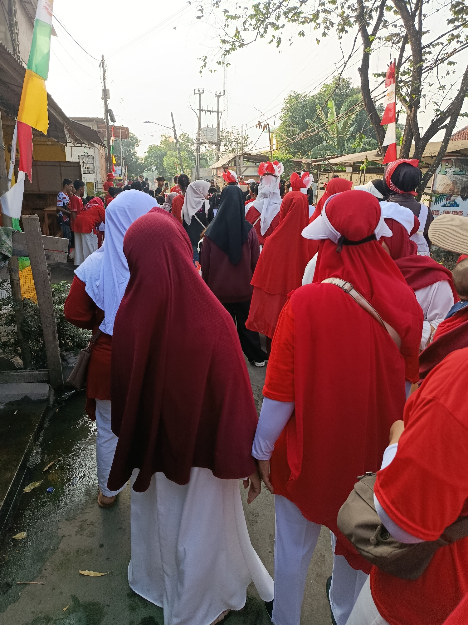 In Balaraja, Indonesia, a street is lined with people, many wearing red and white attire, suggesting a national celebration or event. Flags, including Indonesian flags, are visible, reinforcing this idea. The crowd appears to be moving, possibly in a parade or procession. Buildings and trees line the street, and utility poles with wires are present, indicating an urban or semi-urban environment. The ground is wet, suggesting recent rain or the presence of water. The overall atmosphere seems festive and communal, with people of various ages participating.