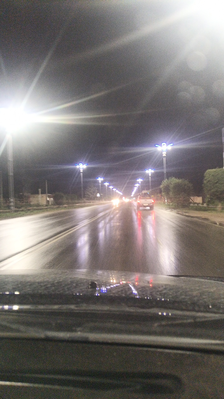 This media file depicts a nighttime view of a wet, multi-lane road in Balykchy, Kyrgyzstan. The scene is illuminated by rows of modern streetlights, which have a distinctive glowing blue outline. The road surface is glistening with water, reflecting the lights and the headlights of passing vehicles, suggesting it is raining or has recently rained.
Several vehicles are visible on the road. In the foreground, the dashboard of a car is partially visible, with water droplets on its surface. Further down the road, a white pickup truck is moving away from the viewer, its brake lights illuminated in red. Other vehicles are seen in the distance, their headlights creating streaks of light on the wet pavement.
The overall atmosphere is one of a wet, dark night, possibly with fog or haze contributing to the diffused lighting. The sky is dark and indistinct, with bright light flares emanating from the streetlights, indicating a long exposure or a bright light source. The setting appears to be a somewhat urban or suburban area, given the presence of well-maintained streetlights and multi-lane roads. There is no visible text in the image. The scene evokes a sense of travel during inclement weather.