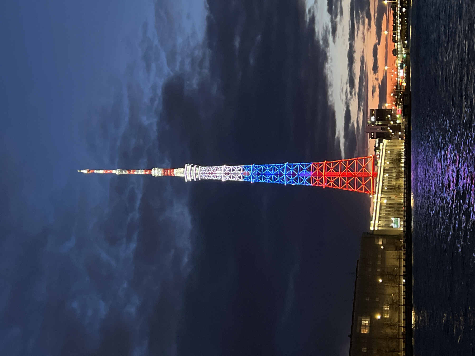 A vertical photograph captures the Saint Petersburg TV Tower brilliantly illuminated in red, white, and blue at dusk or early night. The tall, slender lattice structure of the tower dominates the left side of the frame, with its patriotic lighting scheme – red at the base, white in the middle, and blue towards the top – clearly visible against the darkening sky.
The setting is an urban waterfront in Saint Petersburg, Russia. The sky above the city is a deep, cloudy blue, transitioning to a lighter grey and hints of orange-pink on the horizon where the last vestiges of sunset are visible. Along the opposite bank of a body of water, a cityscape stretches out, featuring numerous buildings with warm, illuminated windows and streetlights that dot the skyline.
The water, likely a river or canal, appears on the right side of the image, reflecting the urban lights and the muted colors of the twilight sky. The surface of the water shows subtle ripples, contributing to the serene atmosphere. There are no people visible, and no discernible text is present in the image. The scene conveys a sense of quiet urban grandeur and patriotic display through the tower's striking illumination.