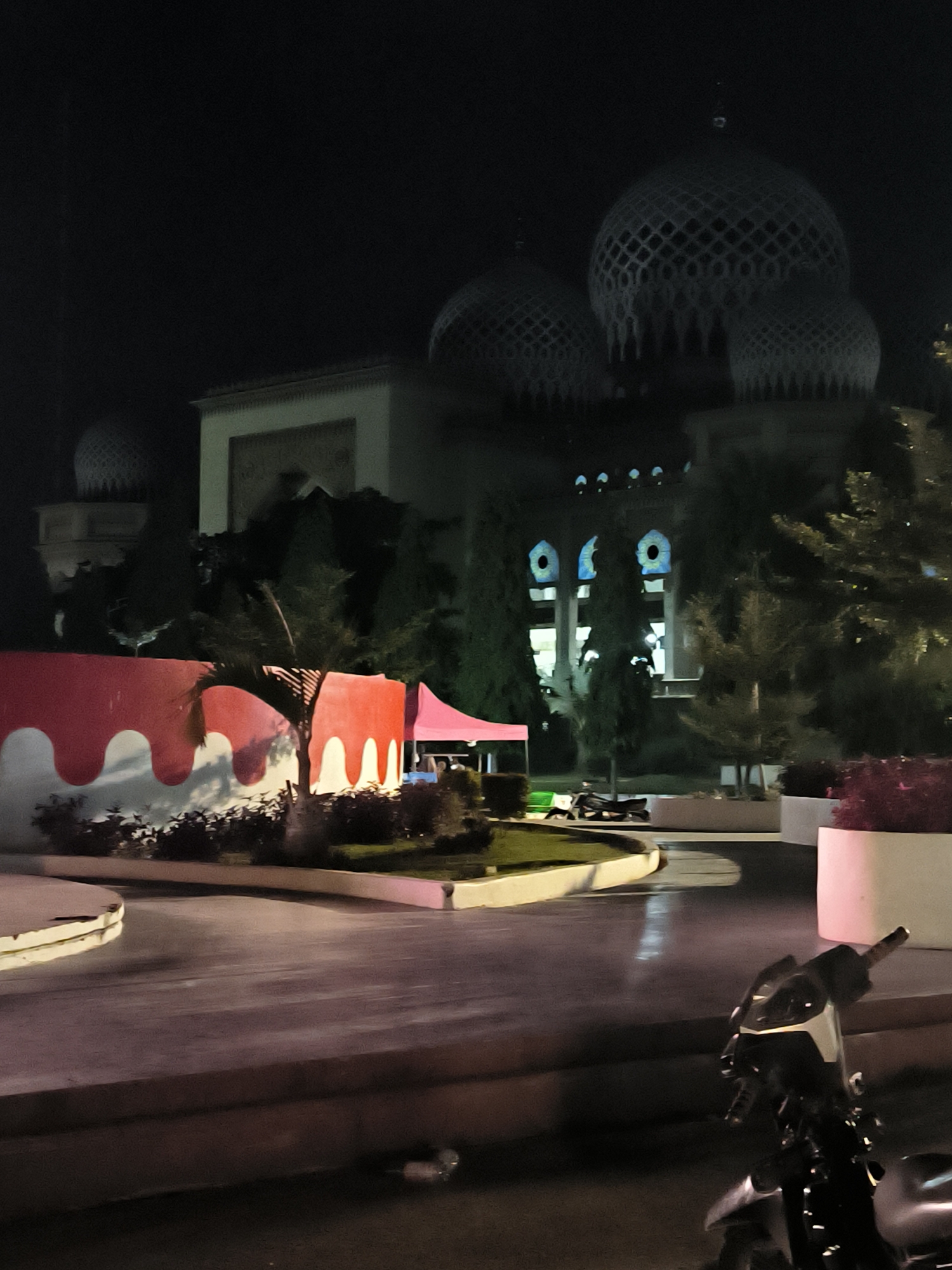 This is a night-time, medium-shot photograph of the Baiturrahman Grand Mosque in Lhokseumawe, Indonesia. The mosque's prominent domes, illuminated by exterior lights, dominate the background. In the foreground, a red and white curved wall borders a garden area. Beyond this wall, a pink tent is visible. A paved area with reflections from streetlights stretches towards the foreground, where a parked motorcycle is partially visible on the right side of the frame. The scene is dark, with the mosque and some landscaping being the primary illuminated elements.