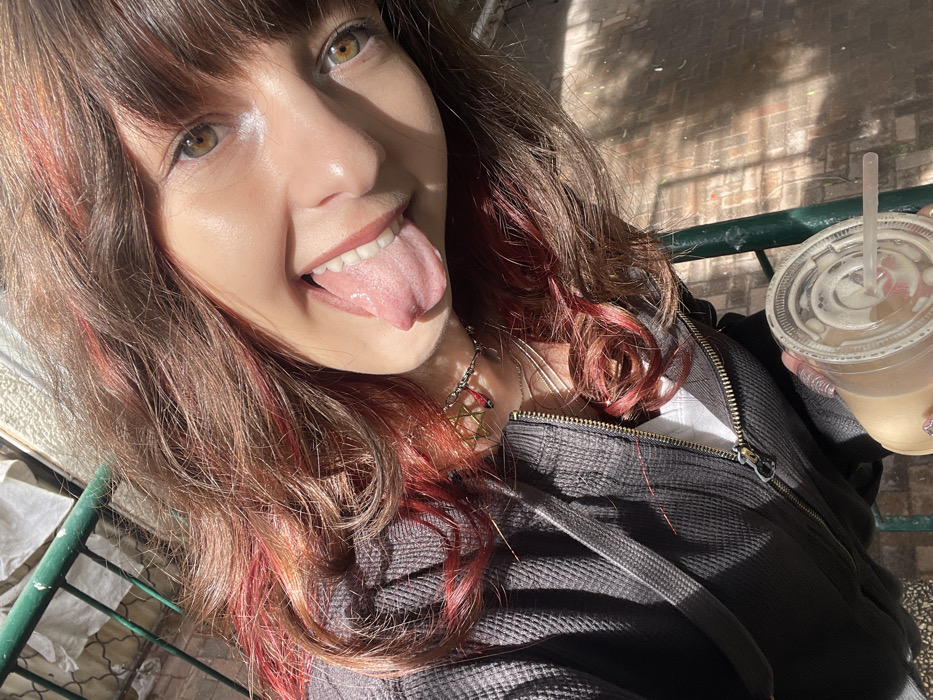 A single individual is depicted in an outdoor, brightly lit setting in Raanana, Israel, captured at an upward-angled perspective. The subject presents with brown hair featuring prominent red highlights and bangs, and possesses green-brown eyes. They are attired in a dark grey, waffle-knit, zippered hoodie worn over a white undershirt. Visible accessories include a silver-colored chain necklace with a Star of David pendant and a small red charm, complemented by an additional thin silver necklace. The subject's left hand, adorned with clear artificial nails, holds a clear plastic cup containing a light brown iced beverage with a lid and straw. The individual is looking directly towards the camera, with their tongue extended. The background elements include a green metal railing, blurry light-colored structures, and a paved or brick-like surface, all under conditions of strong sunlight creating distinct shadows.