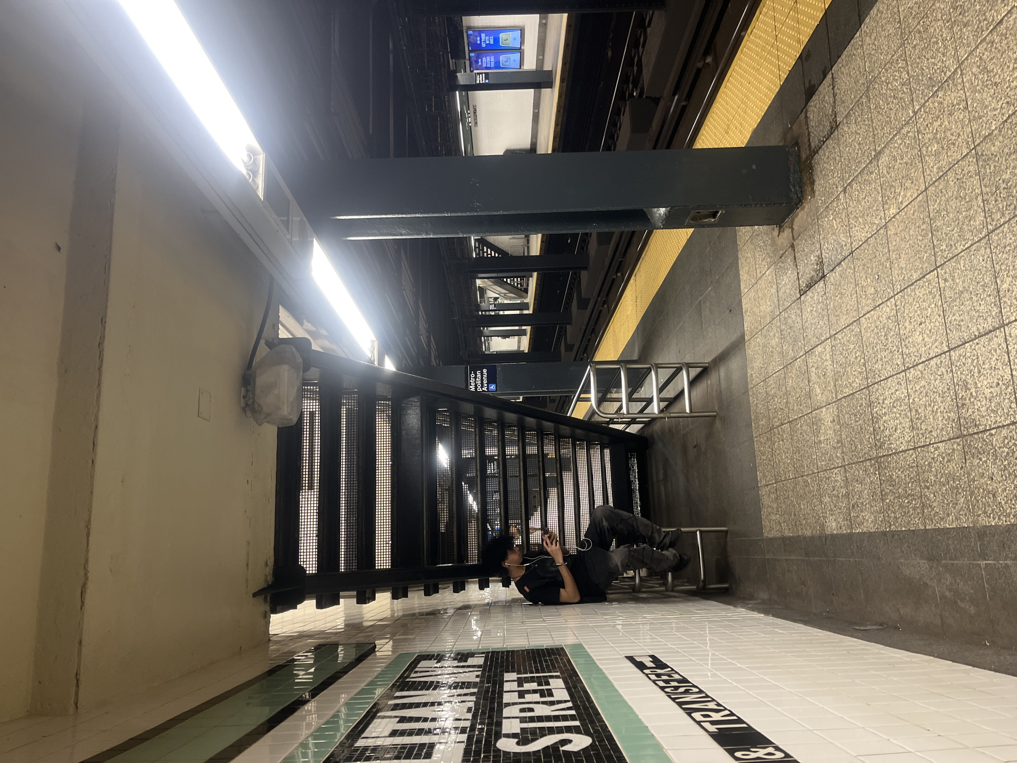 The image depicts an indoor scene, likely a subway station in New York City, United States of America, as indicated by the "Metropolitan Avenue" text on a station sign. The orientation of the image is rotated, showing a person lying on the ground next to a set of stairs.
The main subject is a young person, dressed in dark clothing, with dark, curly hair. They are lying on their back on the white tiled floor of the subway platform, their legs bent. The person is looking at and holding a smartphone, and appears to be wearing earbuds, suggesting they are listening to something or engaged with their device. Their posture and the earbuds suggest a relaxed or perhaps solitary moment.
The setting is a subway platform with prominent features including a black metal staircase. The floor is tiled with white squares, and there are black and green mosaic tiles with text that is partially visible but appears to read "TRANSFER". A yellow tactile warning strip runs along the edge of the platform. Overhead, there are fluorescent lights and industrial-looking metal structures, common in subway environments. The walls are a mix of tiled and painted surfaces. The overall impression is of a functional, utilitarian urban space.
The time of day is not definitively discernible, but the bright overhead lights suggest it is likely not daytime outdoors, or the station is heavily illuminated. The weather is irrelevant as it is an indoor setting. The scene evokes a sense of urban transience and individual engagement within a public space.