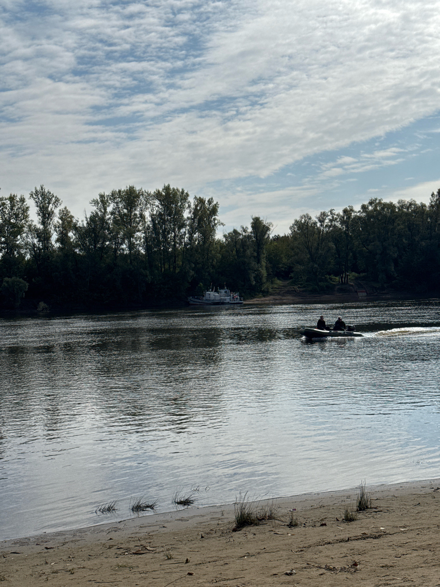 This image depicts a wide river under a cloudy sky. A sandy embankment with sparse vegetation is in the foreground. On the river, a dark inflatable boat with two occupants is moving from left to right, leaving a wake. In the background, a line of dense trees forms the opposite bank. A white boat is also visible docked on the far bank. The scene appears to be a natural landscape, possibly on the outskirts of a city. Given the context, this scene is likely the Belaya River in Ufa, Russia, a popular spot for recreation and natural beauty.