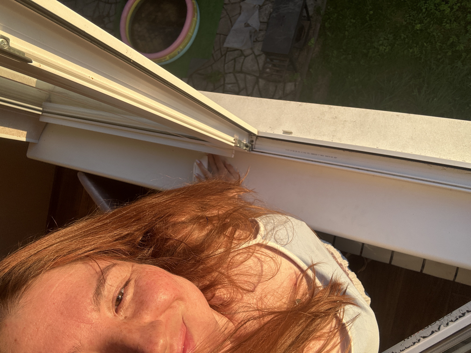 The media file shows a close-up, angled shot of a person with red hair. The person is looking towards the camera, with their face partially visible. They appear to be leaning out of a window, suggesting they are indoors looking out. The setting visible through the open window appears to be an outdoor area with greenery, stone paving, and a circular, inflatable pool with colorful rings. The light suggests it is either early morning or late afternoon, with the sun casting a warm glow. The overall mood seems relaxed and casual. There is no visible text in the media file.