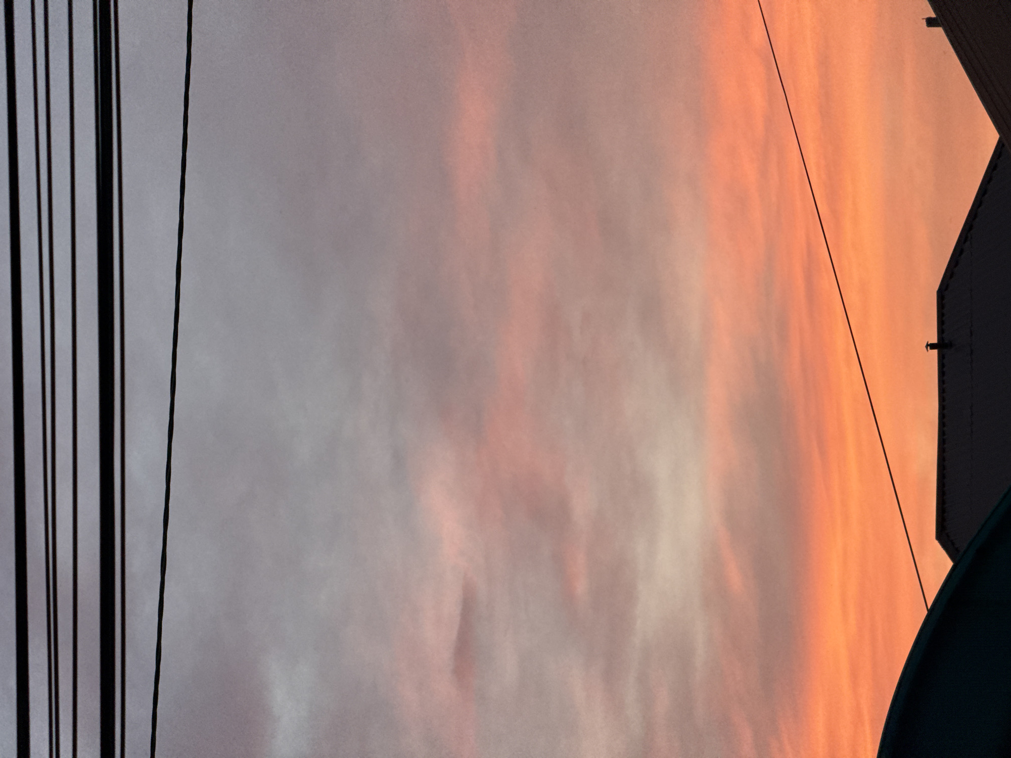 An image captures a sunset over Mikhaylovsk, Russia. The sky is a canvas of vibrant colors, transitioning from a deep orange at the horizon to soft pinks and purples higher up, with hints of grey near the top. Thin, dark power lines crisscross the sky, adding a geometric element to the natural beauty. In the foreground, silhouetted rooftops of houses are visible, their dark shapes contrasting with the illuminated sky. A dark, curved awning also features in the bottom left corner. The overall scene evokes a sense of calm and the quiet beauty of an evening in a Russian city.