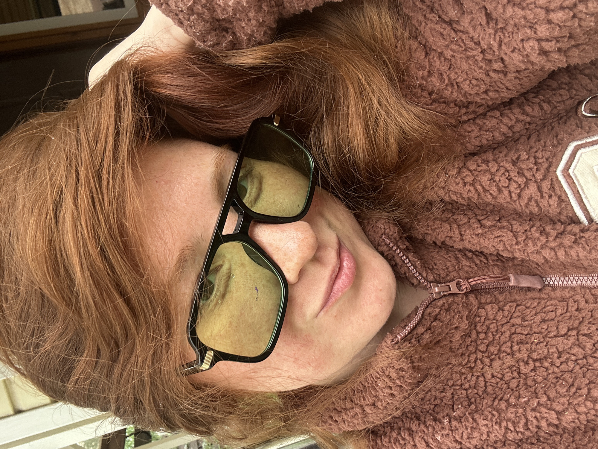 A person with red hair is wearing sunglasses and a brown, zippered, sherpa-style jacket. The photograph is taken from an elevated perspective, looking down at the person. The background is slightly visible and blurry. It is impossible to tell with certainty if the image was taken in Nizhny Novgorod, Russia, due to the lack of context and recognizable landmarks.