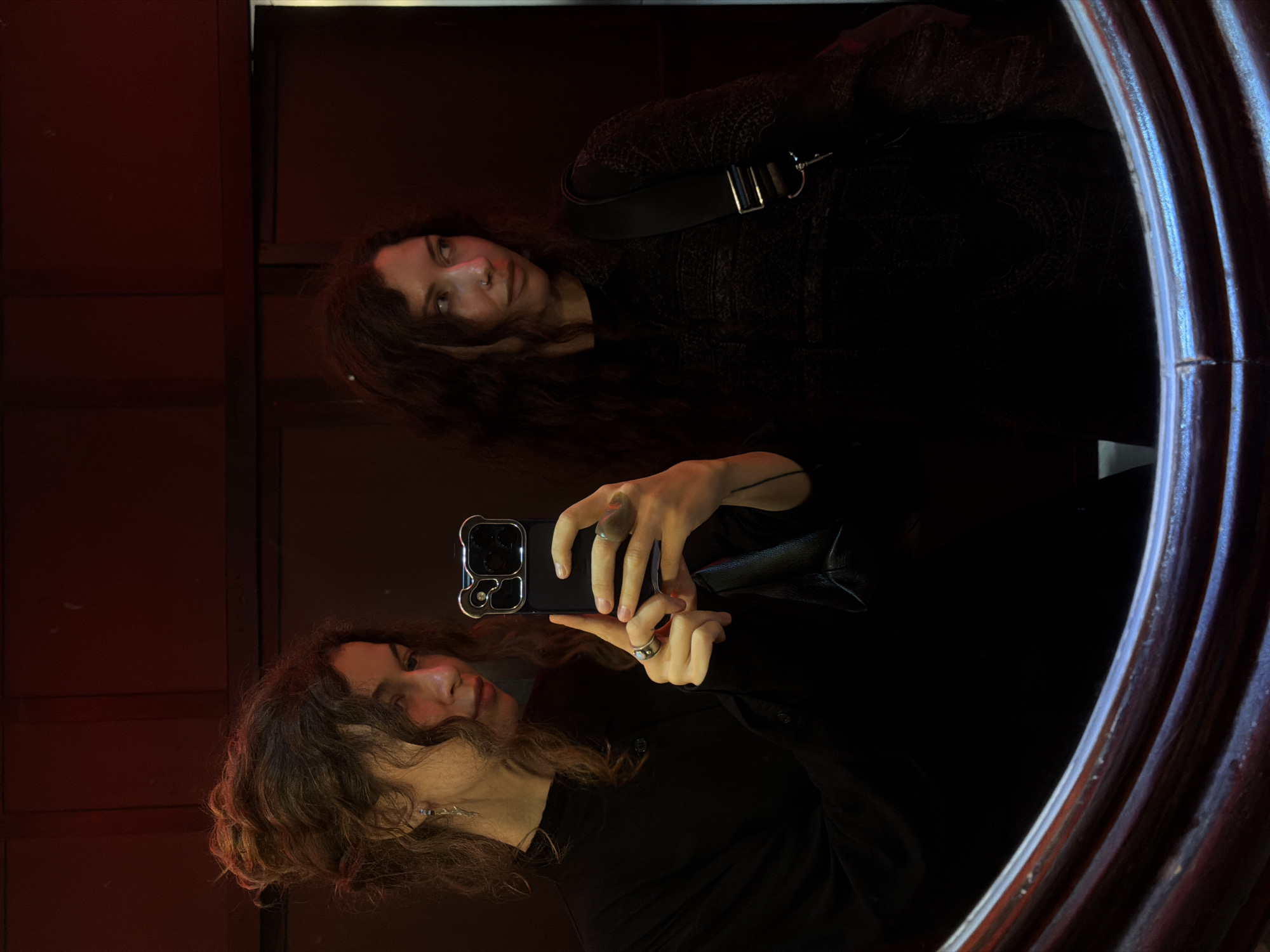 This image captures a mirror selfie featuring two individuals reflected, or possibly one person reflected twice, in a dimly lit indoor setting. The overall aesthetic is dark and moody, with warm red and orange light illuminating parts of the scene from the left, possibly from ambient lighting or wall color.
In the foreground, a person with medium-length, wavy dark brown hair is holding a smartphone (likely an iPhone, identifiable by its camera array design) to take the selfie. They are wearing a dark, long-sleeved top and have a silver-colored ring on their right index finger. Their expression is neutral to slightly smiling.
Further back and higher in the mirror's reflection is a second individual. This person also has dark, wavy hair and is wearing a dark, possibly textured or patterned, long-sleeved garment. A shoulder bag with a prominent rectangular metal buckle is slung across their body. This individual appears to have a nose piercing and a neutral or contemplative expression.
The mirror itself has a dark, possibly ornate wooden frame, visible on the right side of the image. The background behind the reflections is predominantly dark, suggesting a room or corridor with low lighting or dark-colored walls. No discernible text is visible within the image. The photo depicts a casual moment captured in a low-light environment, taking place in Đà Nẵng, Vietnam.