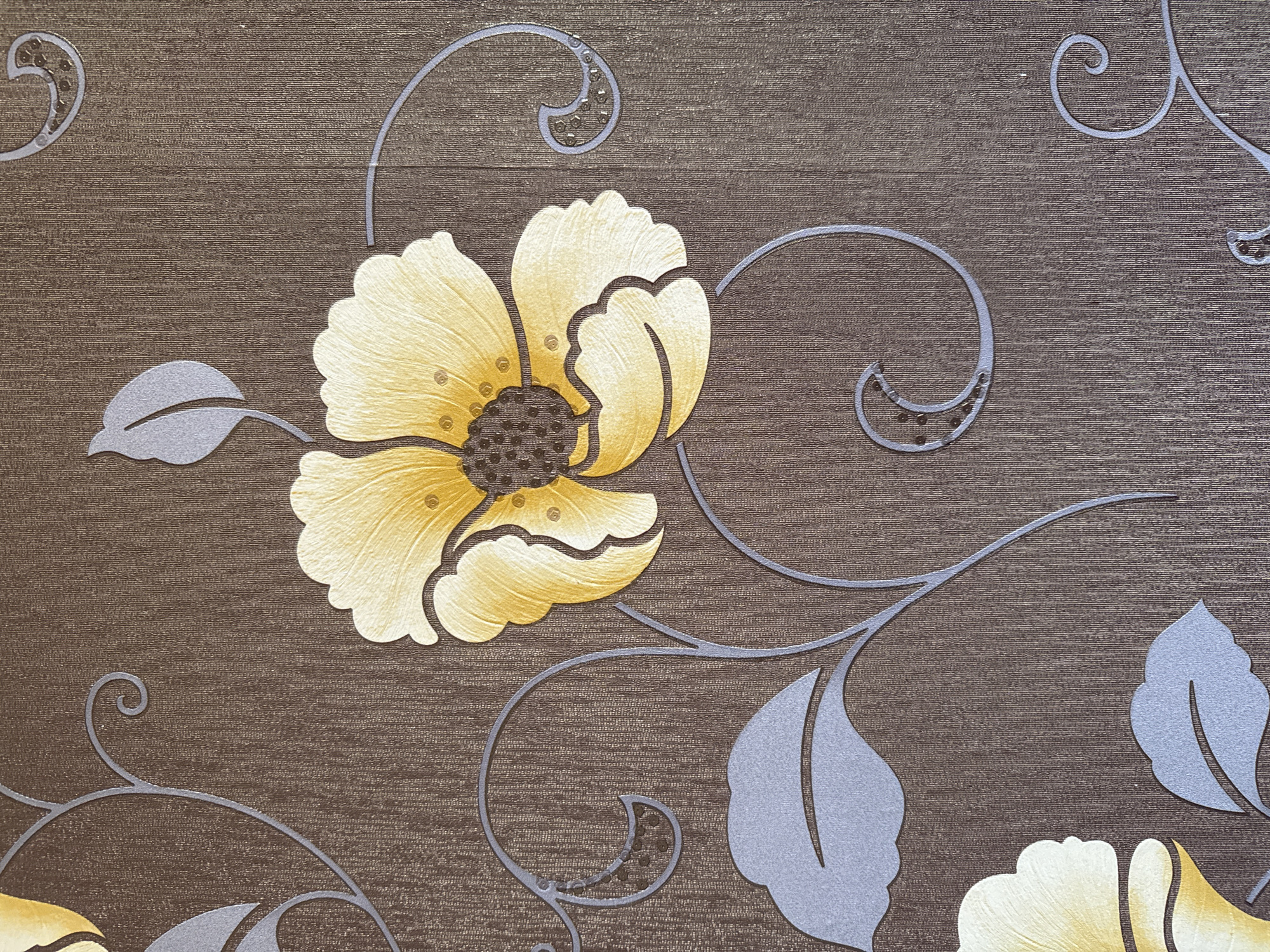 The image is a close-up, vertical shot of floral wallpaper with a brown textured background. The wallpaper features stylized yellow flowers with dark brown centers, interspersed with light gray leaves and swirling vine-like patterns. The floral motifs and accompanying decorative elements are raised from the background, giving the wallpaper a tactile quality. The lighting appears to be uniform, highlighting the texture and design of the wallpaper without casting harsh shadows. There are no people or any indication of a specific location in Bishkek City, Kyrgyzstan. The image focuses solely on the decorative wallpaper pattern.