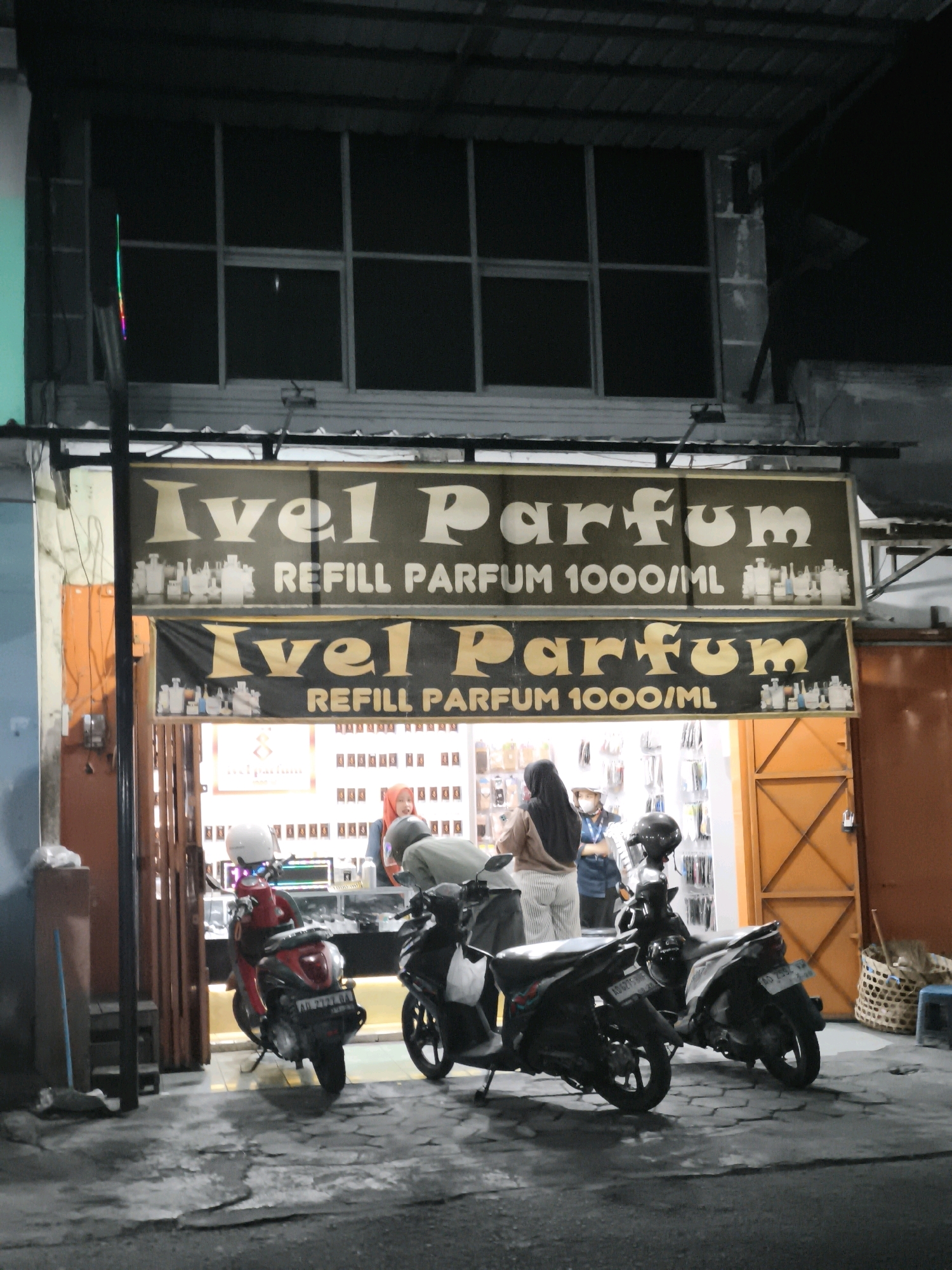 The image depicts the storefront of "Ivel Parfum" in Desa Donohudan, Indonesia, at night. Two signs with the store's name and "REFILL PARFUM 1000/ML" are displayed. Inside the store, shelves are stocked with perfume bottles, and three individuals are visible, two of whom appear to be customers or staff. In front of the store, two motorcycles are parked, one red and one black. The ground is wet, suggesting recent rain. The overall scene is dimly lit, with the store's interior providing the primary light source.