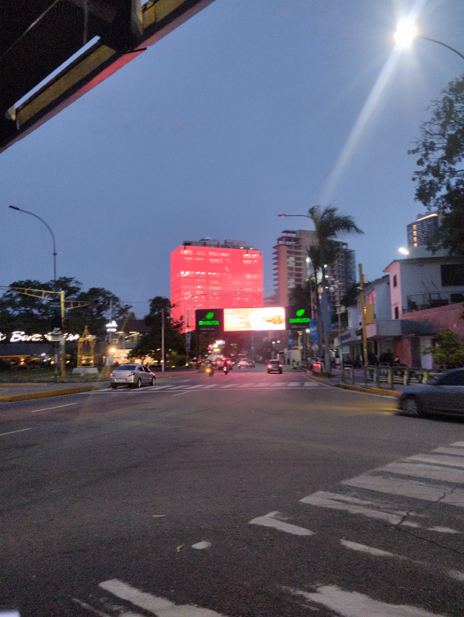 The image depicts a street scene in Caracas, Venezuela, at dusk or dawn. A large red building dominates the view, likely the headquarters of the Banco Mercantil, situated at Avenida Libertador. Streetlights illuminate the area, and there's moderate vehicle traffic. A pedestrian crossing is visible in the foreground. Several buildings are visible in the background. The sky is a gradient of dark blue to lighter shades.