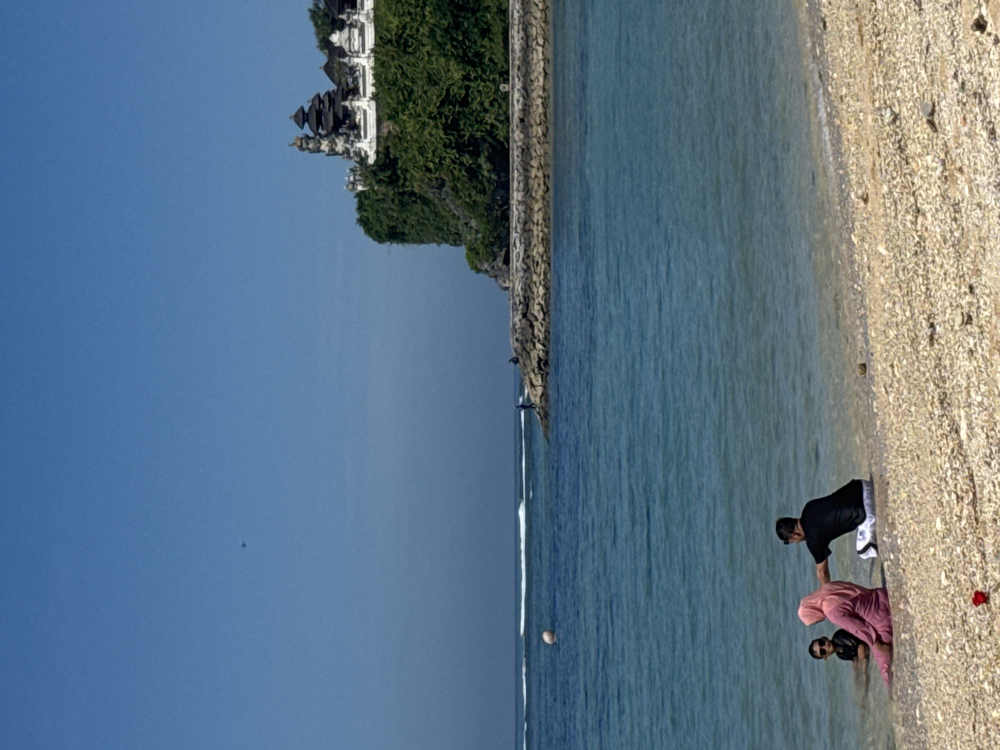 A horizontal outdoor shot depicts a beach scene. On the right side of the image, the sandy beach transitions to the ocean, which fills the majority of the scene. In the lower right, three people are standing in the shallow water near the beach. One person is wearing black and white clothing, while another wears pink clothing. A third person is partially visible, wearing black clothing. On the left side of the image, the clear blue sky is visible. In the upper left, a building is perched on a cliff covered with greenery. A wall divides the greenery from the ocean. A small, dark object, possibly a bird or insect, can be seen in the sky. A light-colored object floats in the ocean near the shoreline. A red object lies on the beach near the people. The location appears to be in the city of Unknown, Unknown.