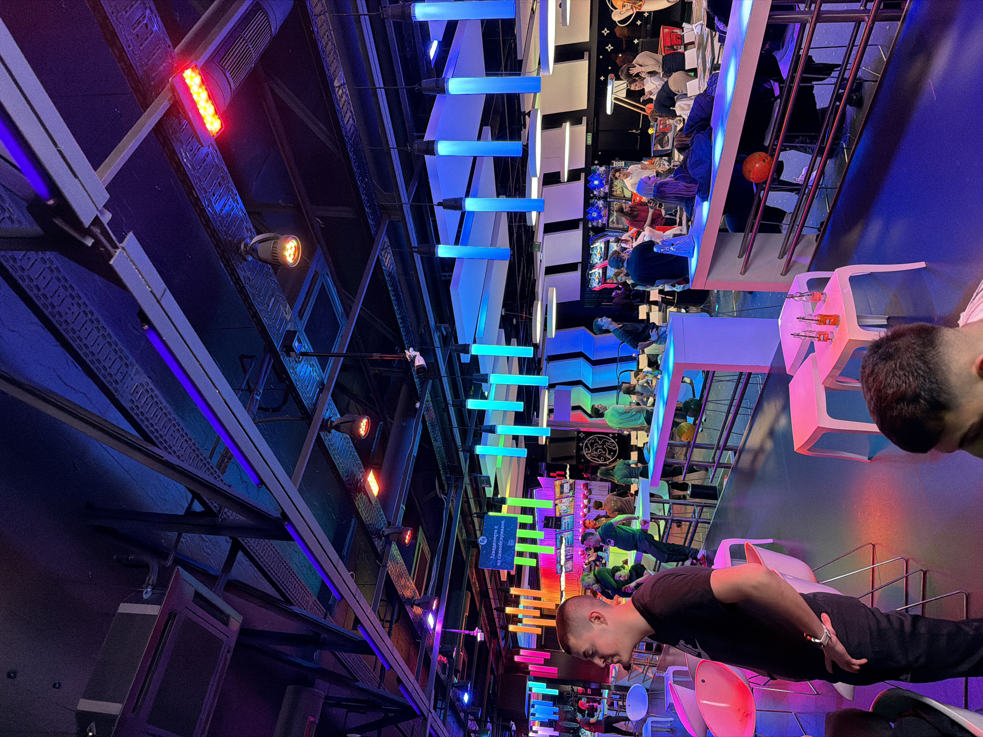 The image is an indoor shot of an entertainment venue, possibly an arcade or a bar, located in Sofia, Bulgaria. The scene is brightly lit with neon lights of varying colors, including blue, purple, orange, and green, arranged in horizontal stripes across the ceiling, along with spotlights. Metal structural beams crisscross the ceiling, contributing to the industrial aesthetic.
A crowded area filled with people engaged in activities is visible. Several are seated at a bar with metal railings, suggesting a leisure environment. Various objects, including food, drinks, and bowling balls, are scattered on the surfaces, hinting at the activities available.
In the foreground, two men are standing near the edge of the frame, partially obscuring the view of the interior. One man is dressed in black attire, and his features are partly visible. The presence of people in the foreground indicates the image's focus on capturing the environment.