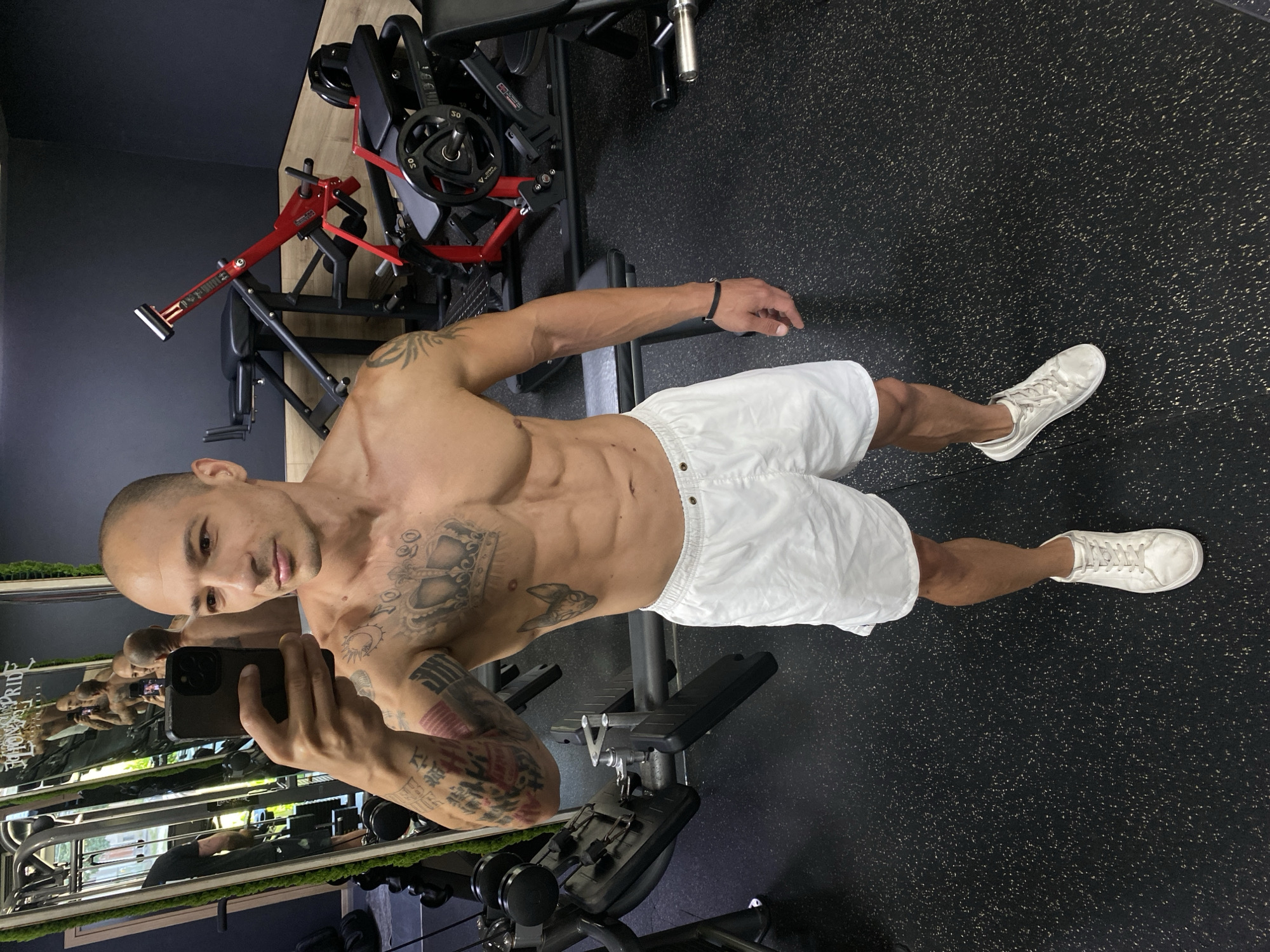 A man with a shaved head and tattoos on his arms and chest stands in a gym, taking a selfie in a mirror. He is wearing white shorts and white sneakers. The gym has black rubber flooring and various weightlifting machines. The mirror reflects the man and other gym equipment, including a leg press machine and weight racks. The background shows a dark wall and wooden accents. The man's physique suggests he is actively engaged in fitness. The overall scene is consistent with a workout session in a modern fitness facility.